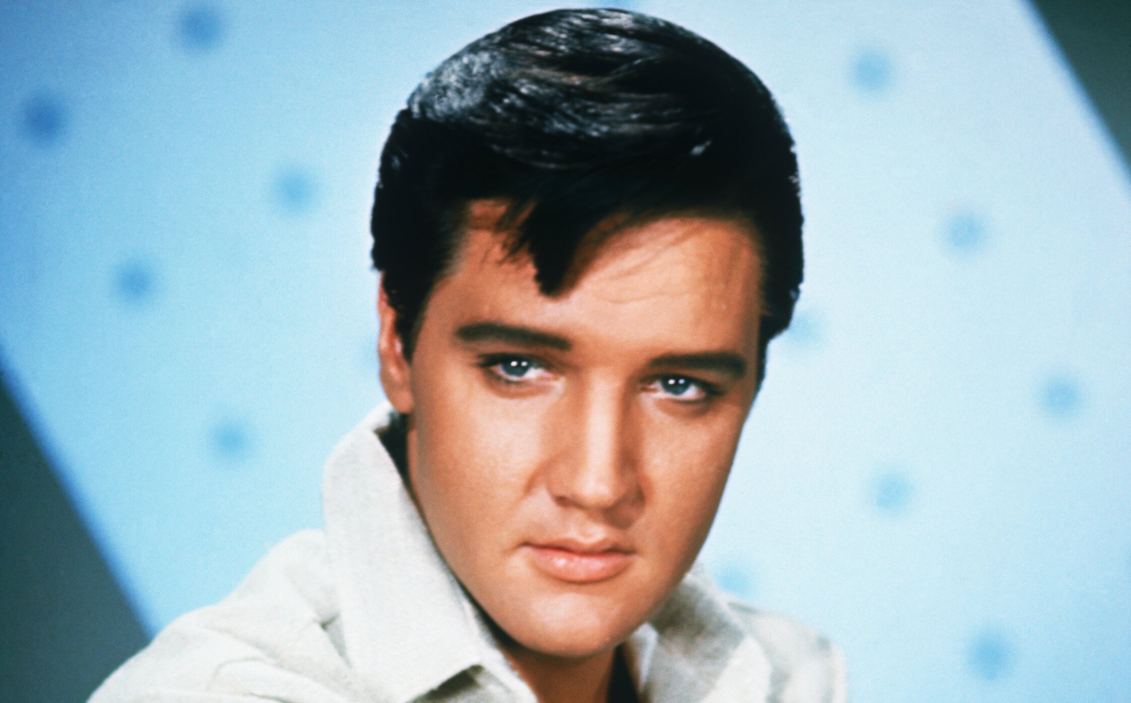 A close-up of Elvis Presley. He's in a white shirt in front of a light blue background.