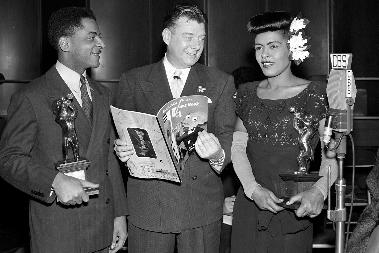 Arthur Godfrey and Teddy Wilson smile at Billie Holiday, who wears a flower in her hair and holds an award