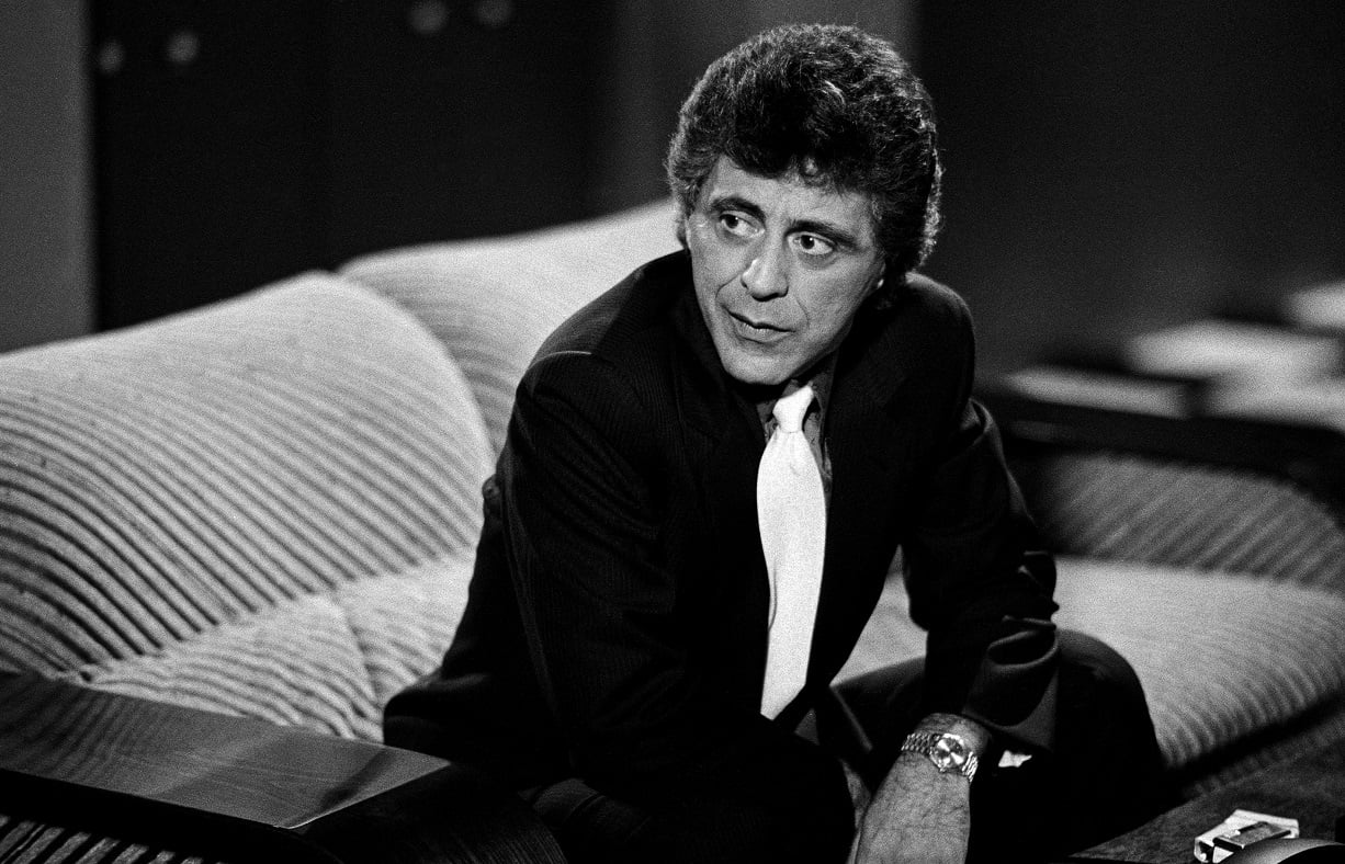 Frankie Valli sits on a couch in a dark suit and white tie as Frank Doss in 'Miami Vice'
