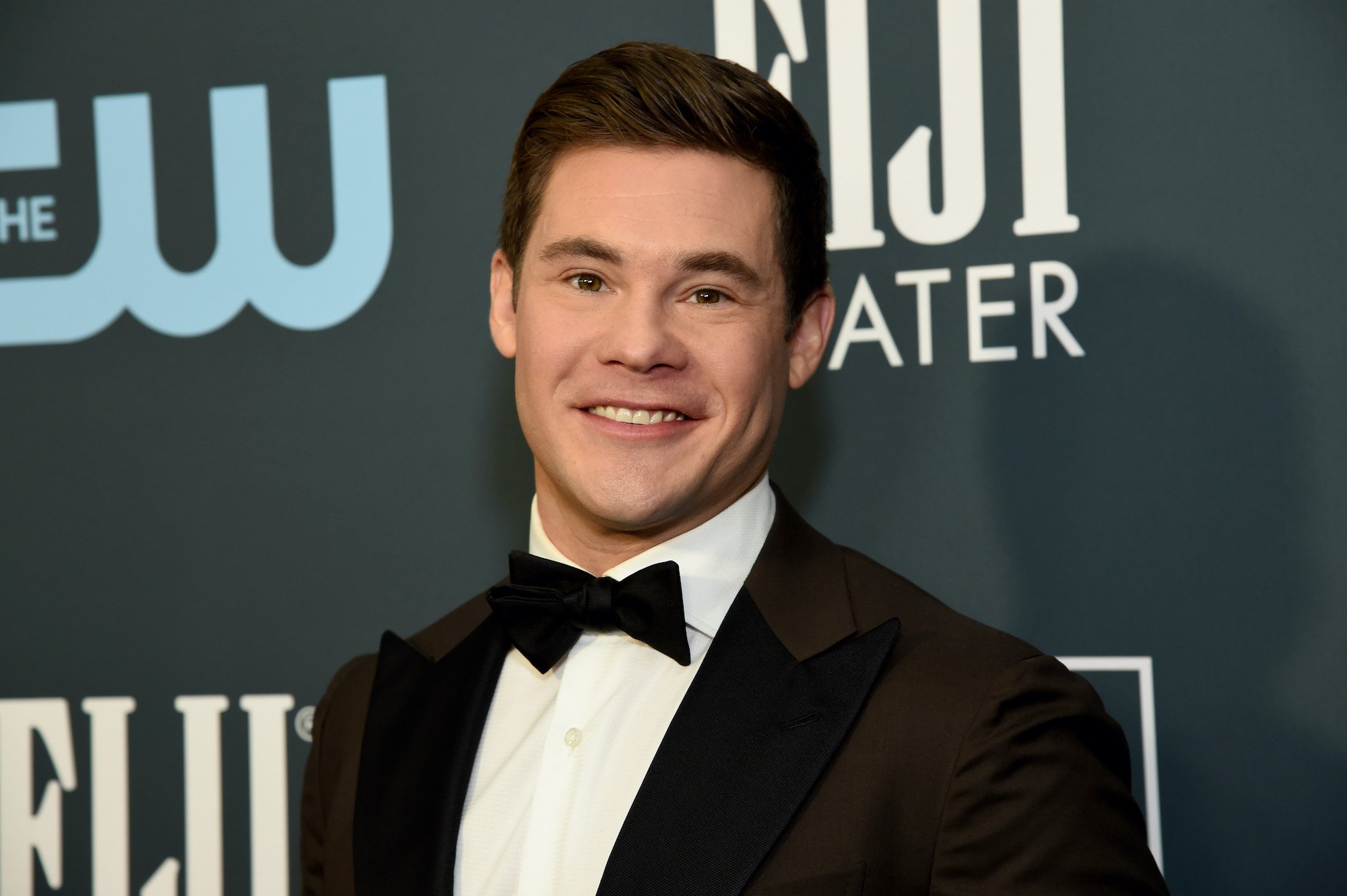 Adam Devine smiling in front of a gray background