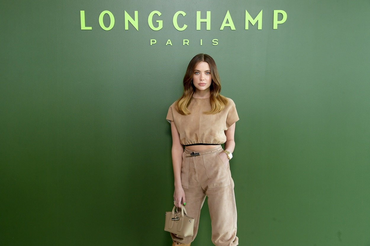 Pretty Little Liars alum Ashley Benson poses under a Longchamp sign wearing a tan outfit and matching purse