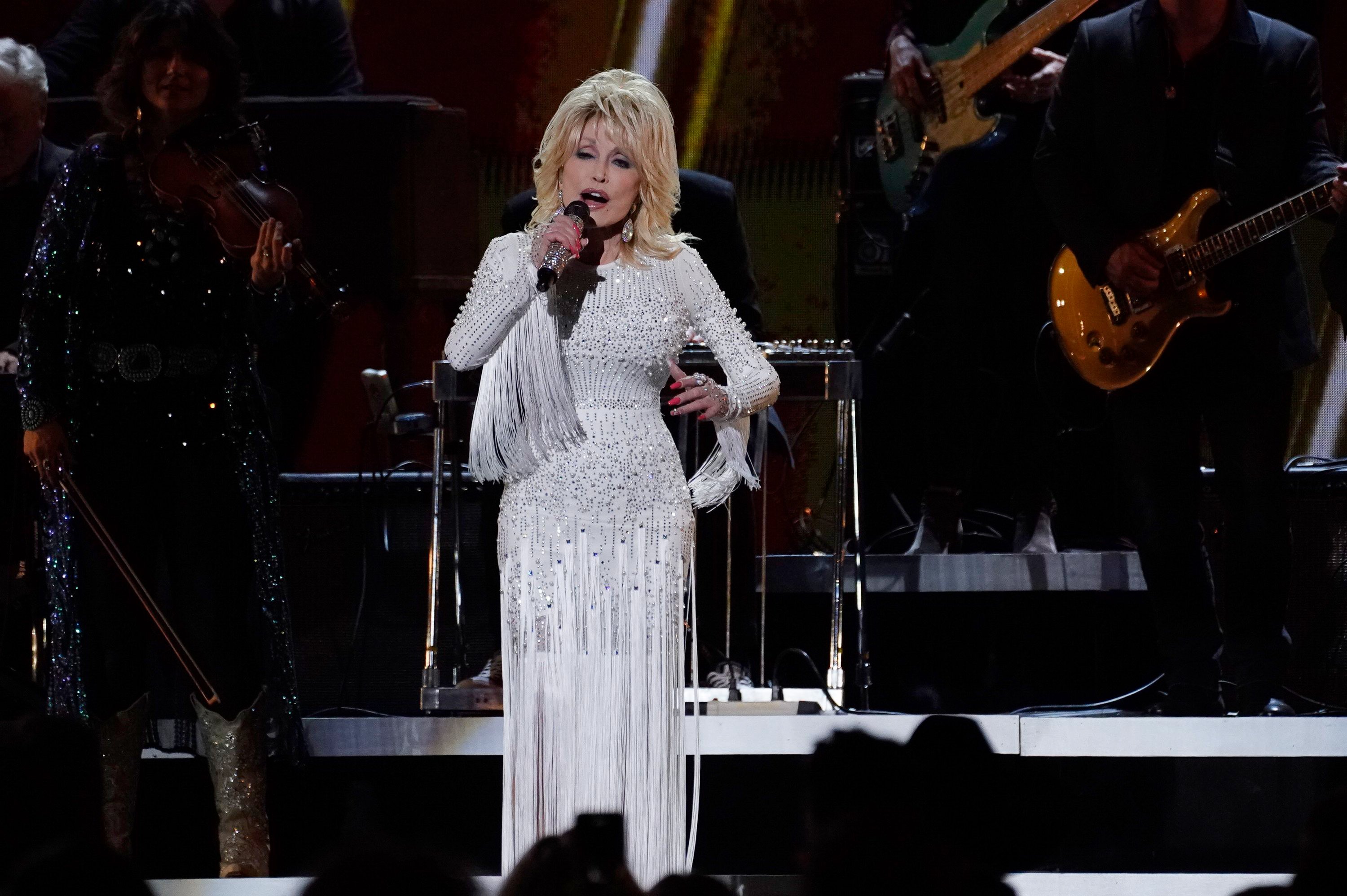 Dolly Parton singing on stage. She's in an all-white fringe dress.