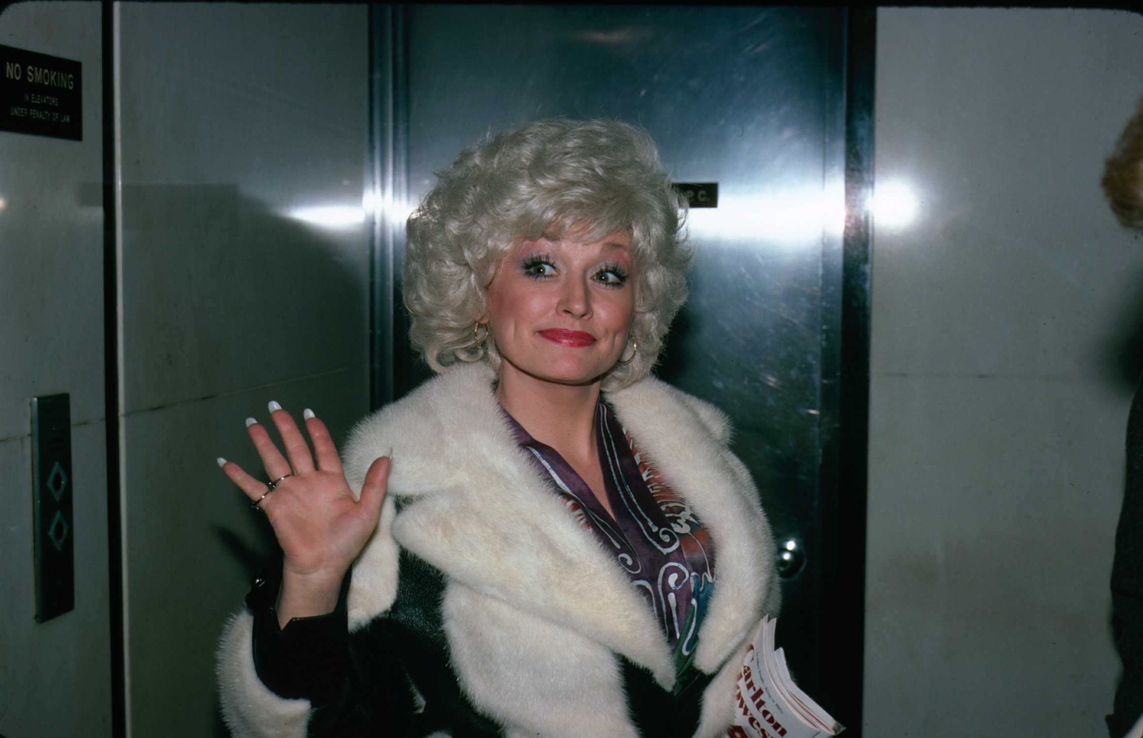 Dolly Parton waves to the camera in a fur coat.