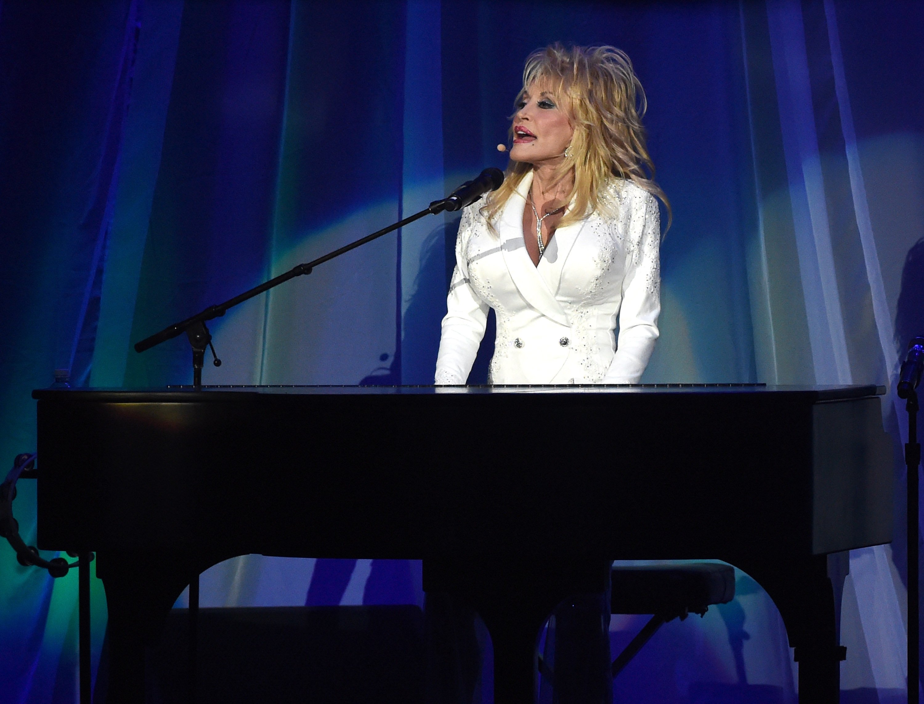 Dolly Parton performing on stage. She is sitting and singing at a piano in an all-white outfit.