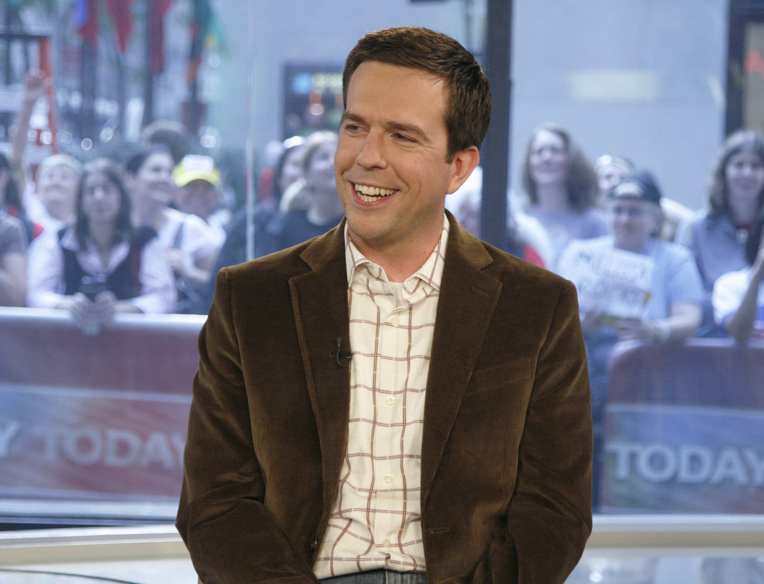 Ed Helms in a brown jacked and plaid shirt, sitting and smiling