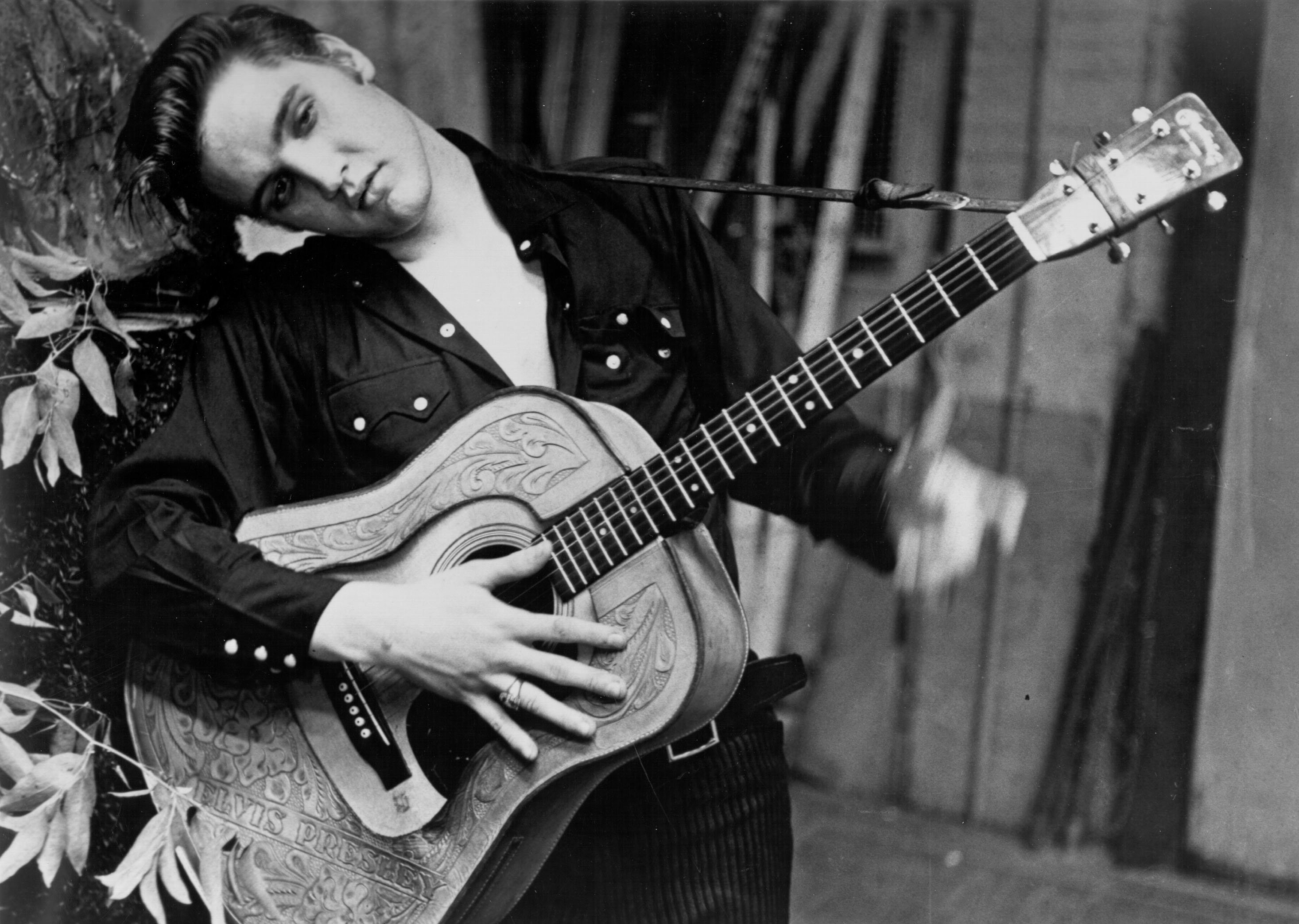 A portrait of Elvis Presley in black and white. He's leaning against a wall, holding his guitar.