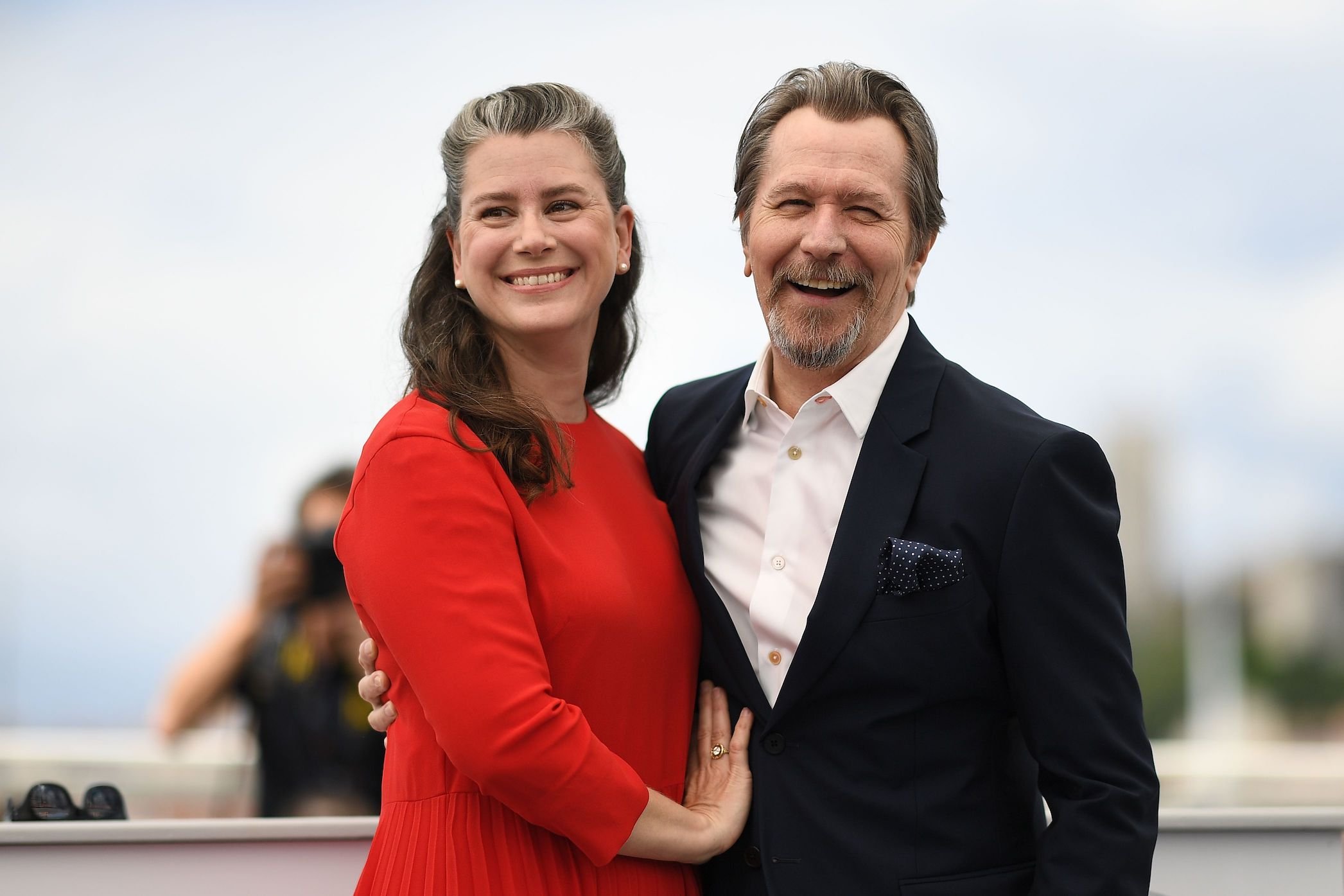 Gary Oldmans' spouse, Gisele Schmidt, standing next to Gary Oldman with her hand on his chest while smiling at the camera