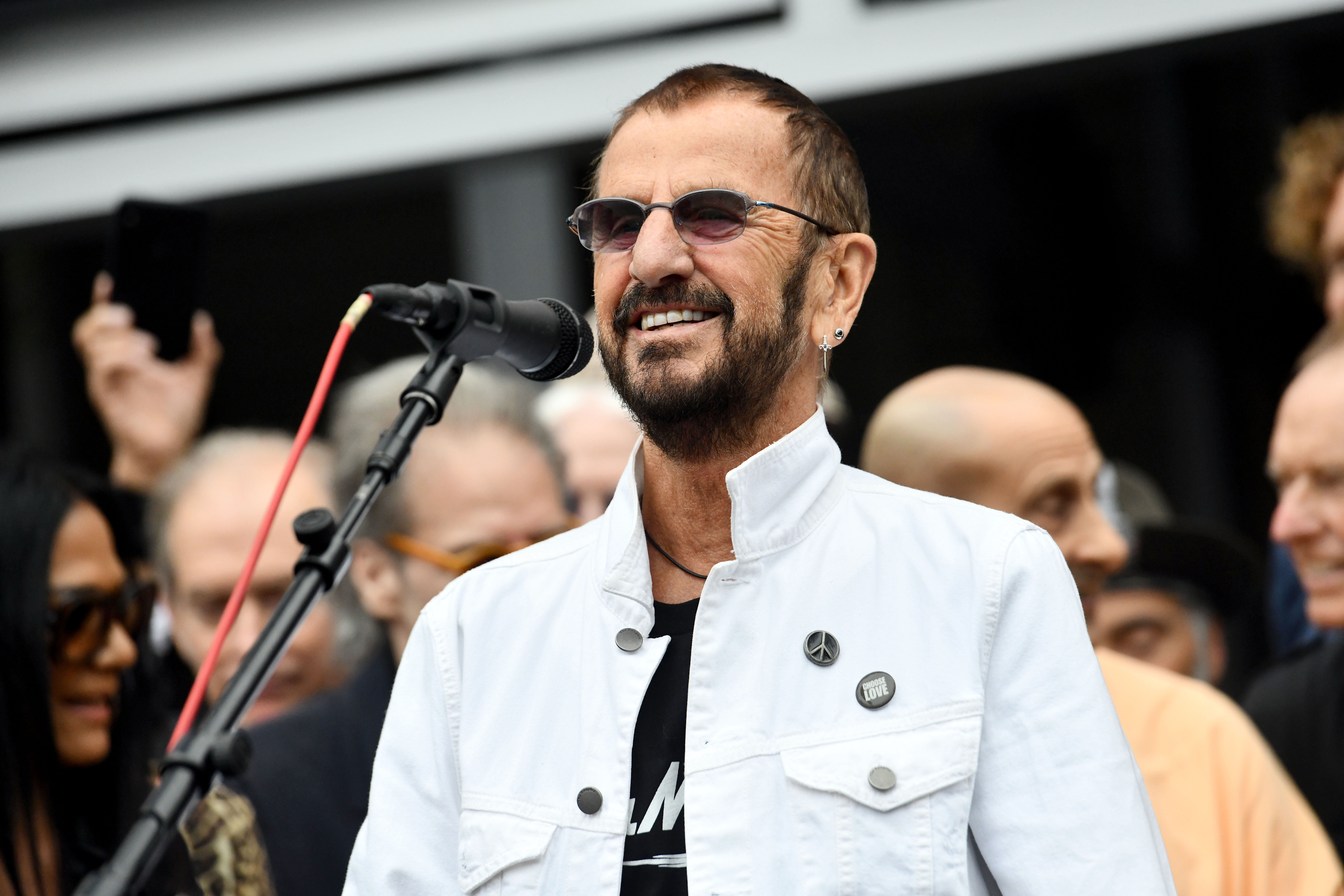 Former Beatle Ringo Starr smiles as he greets a crowd wearing a white jacket and his trademark violet sunglasses.