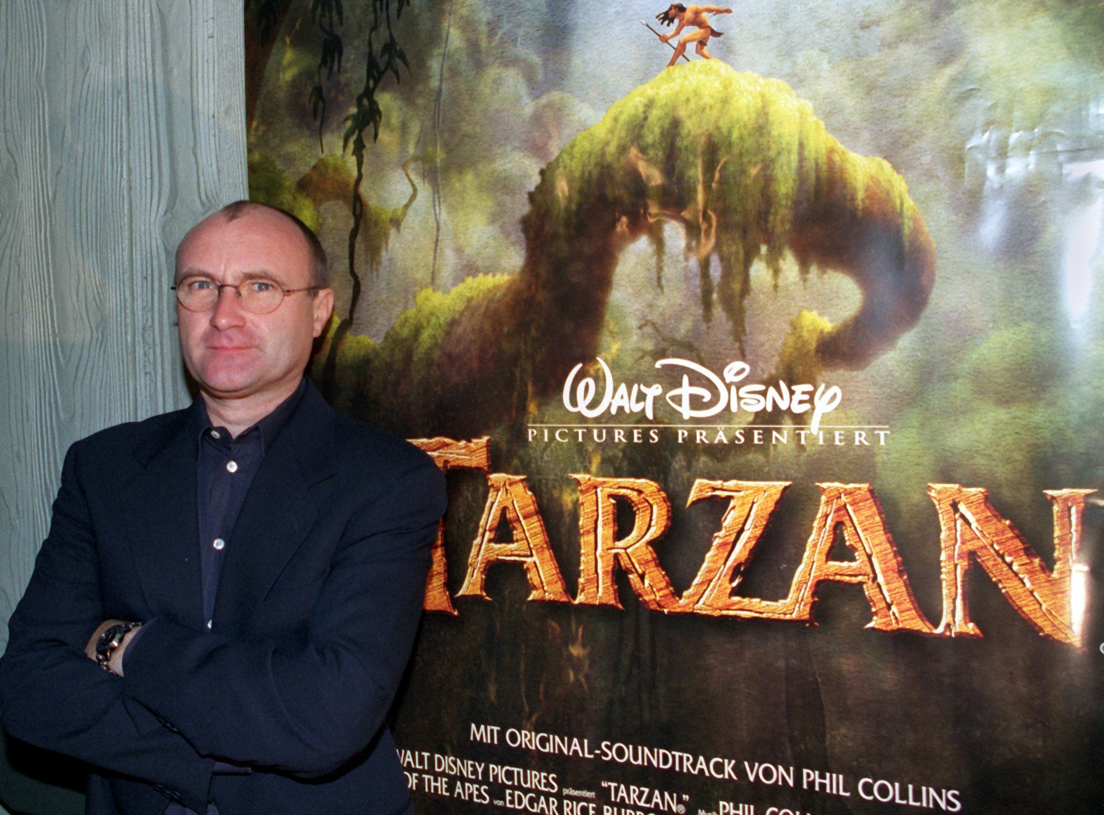 Phil Collins stands in front of a promotional poster for the Disney animated film 'Tarzan,' for which he composed the soundtrack, 1999