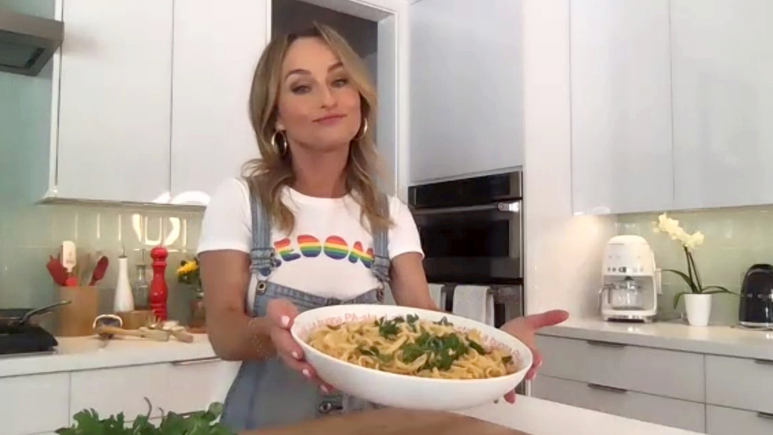 Celebrity chef Giada De Laurentiis stands in her kitchen wearing coveralls and holding a large bowl of pasta.