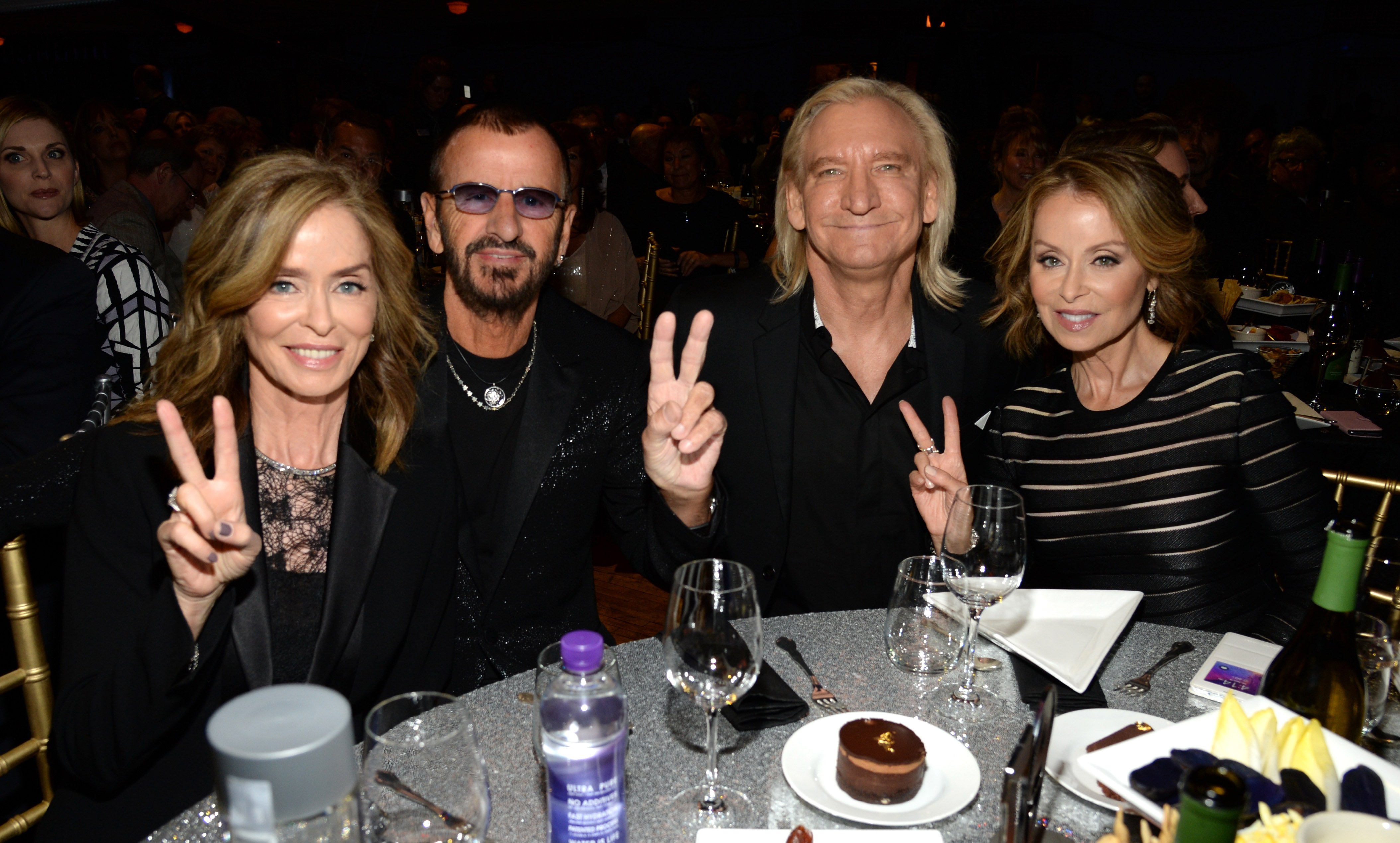 Barbara Bach, Ringo Starr, Joe Walsh, and Marjorie Bach are seated at a table at the 2015 Hall of Fame Induction Ceremony. The group offered peace signs as they posed for a photo.
