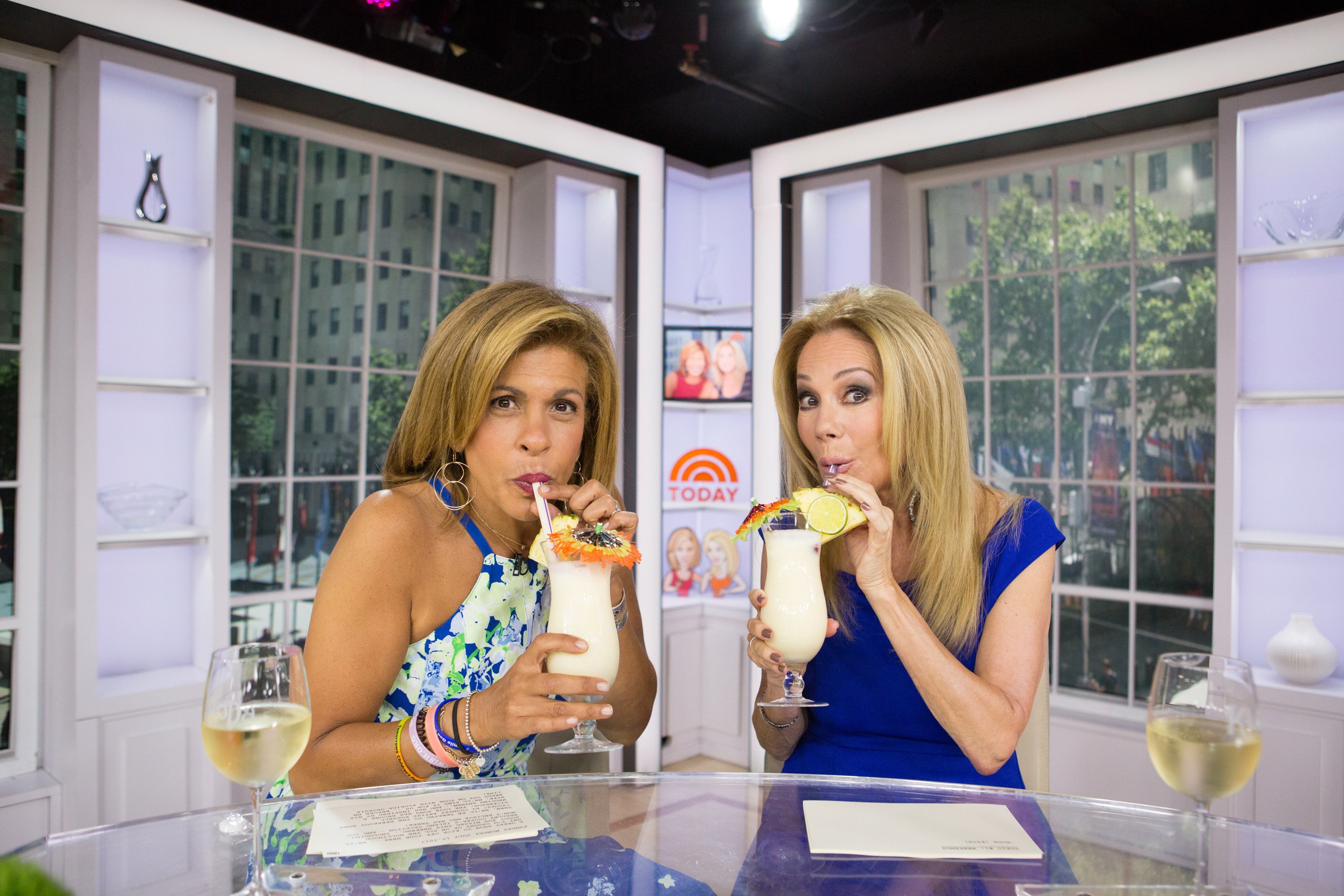 Hoda Kotb in a floral dress and Kathie Lee Gifford in a blue dress, both drinking from straws at the desk of 'Today with Kathie Lee & Hoda' 