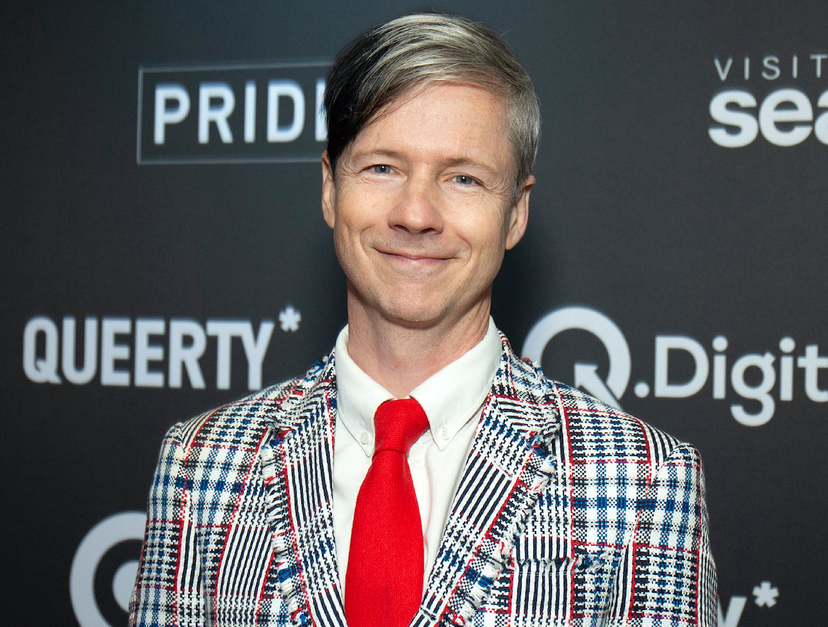 John Cameron Mitchell in a black, white, red, and blue suit, white shirt, and red tie in front of a grey background