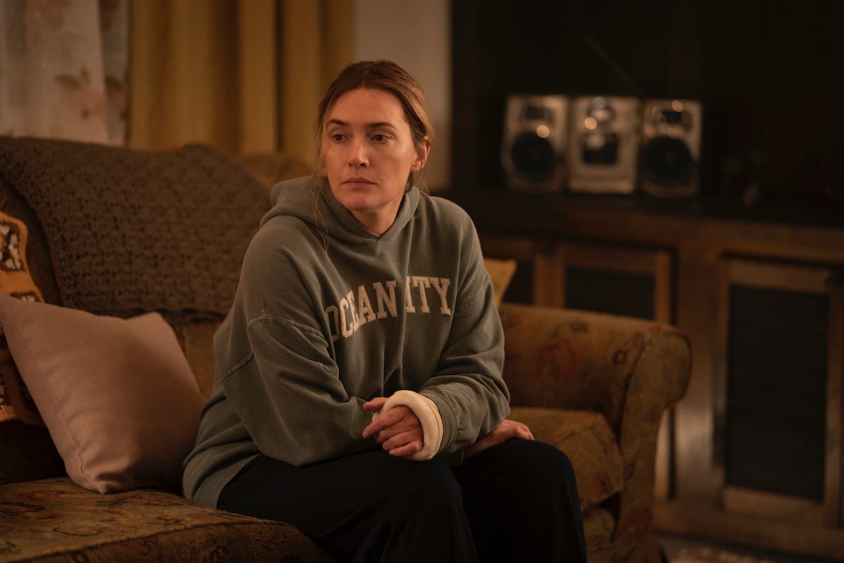 Kate Winslet sits on a couch wearing a hooded sweatshirt during a scene from 'Mare of Easttown'
