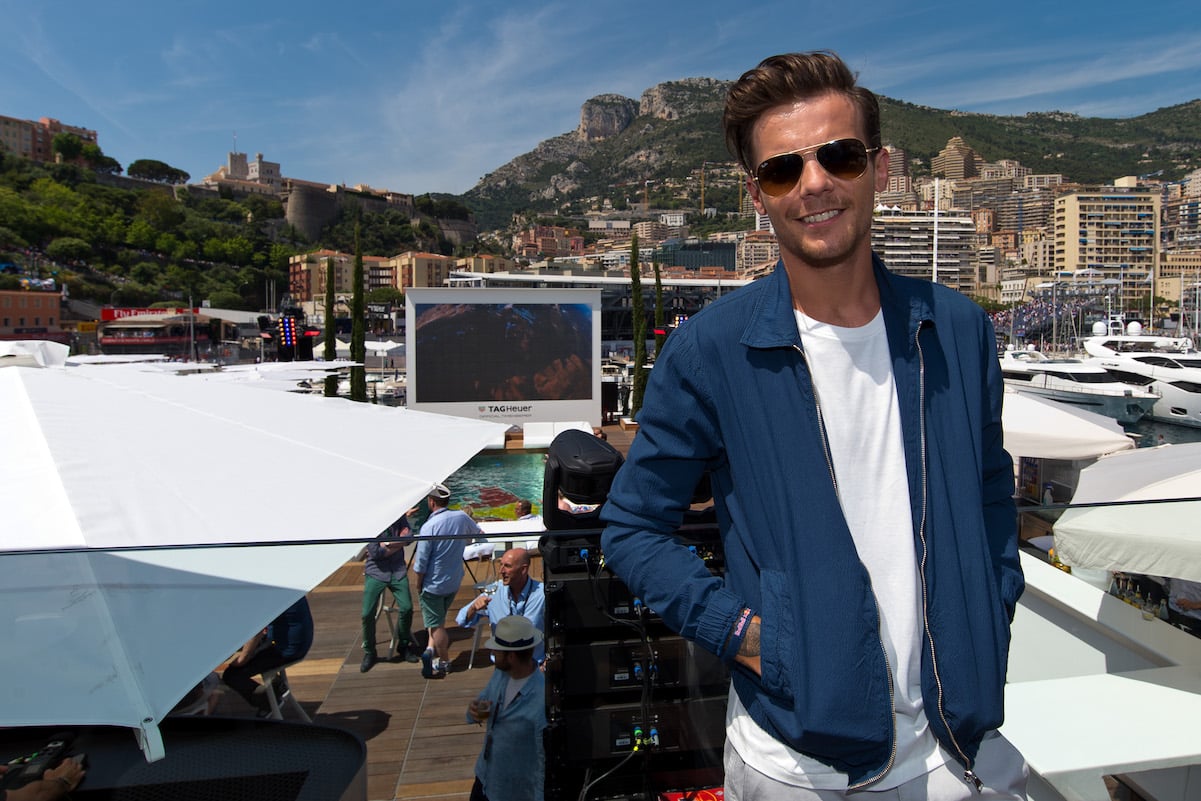 Louis Tomlinson in a blue jacket, white t-shirt, and sunglasses smiling in front of a mountain