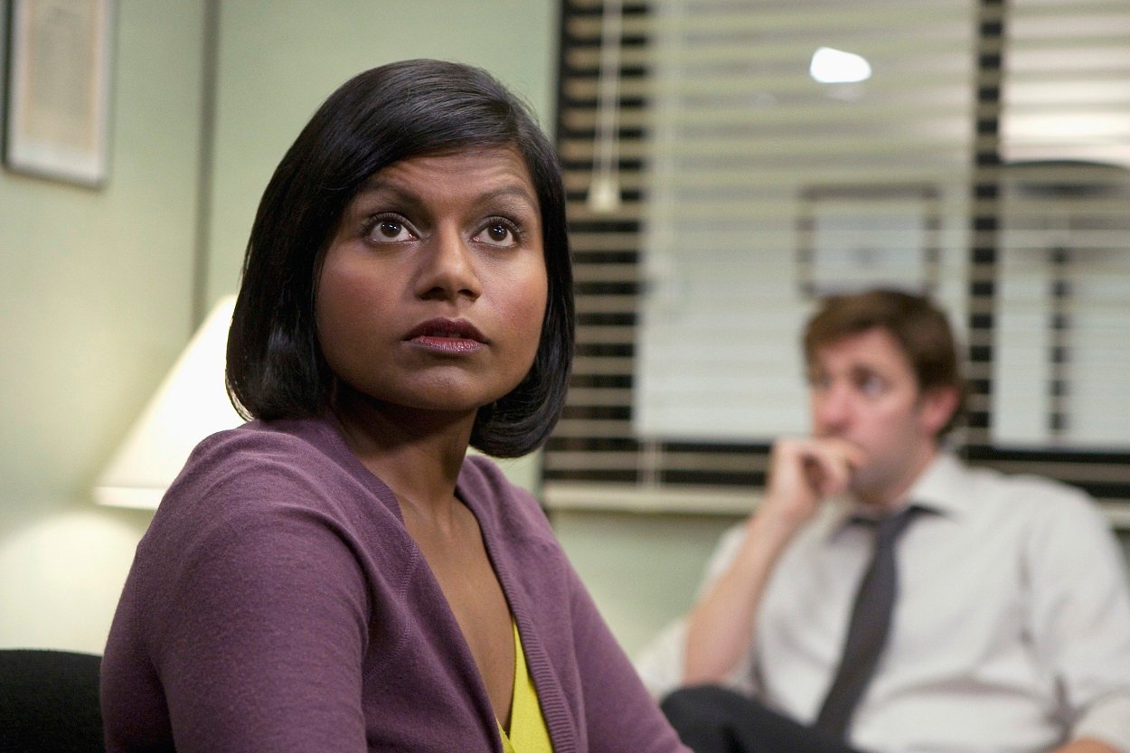 Mindy Kaling films an episode of The Office in a purple sweater. John Krasinski is out of focus in the background