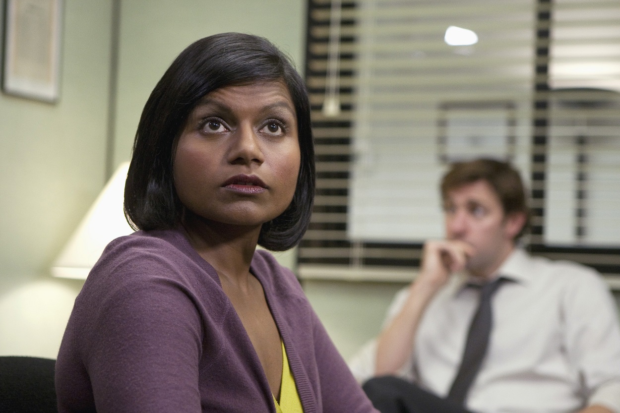 Mindy Kaling films an episode of The Office in a purple sweater. John Krasinski is out of focus in the background