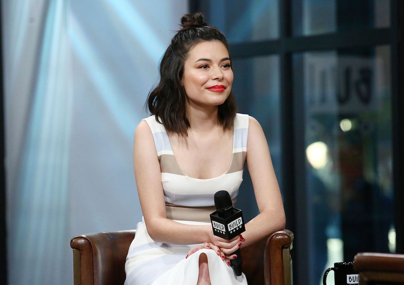 Miranda Cosgrove sitting in white dress, holding microphone