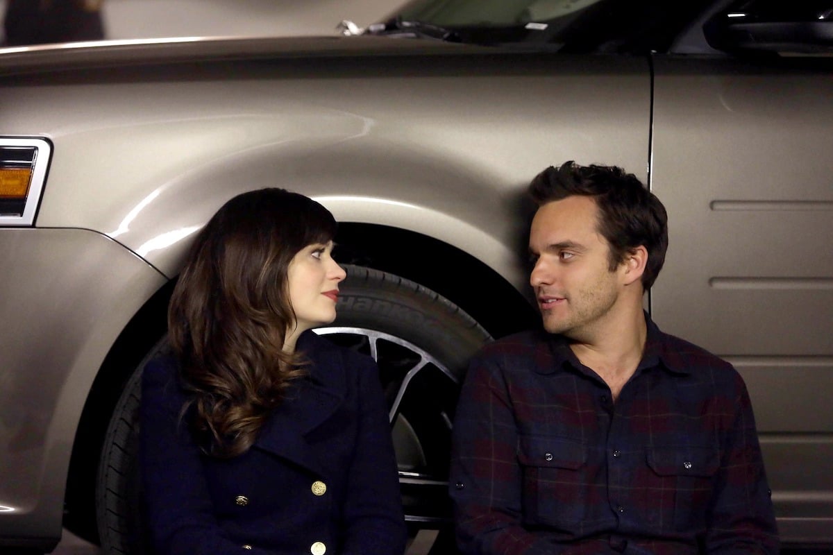 Actors Zooey Deschanel as Jess and Jake Johnson as Nick sit next to each other against a car in a scene on the TV show 'New Girl'