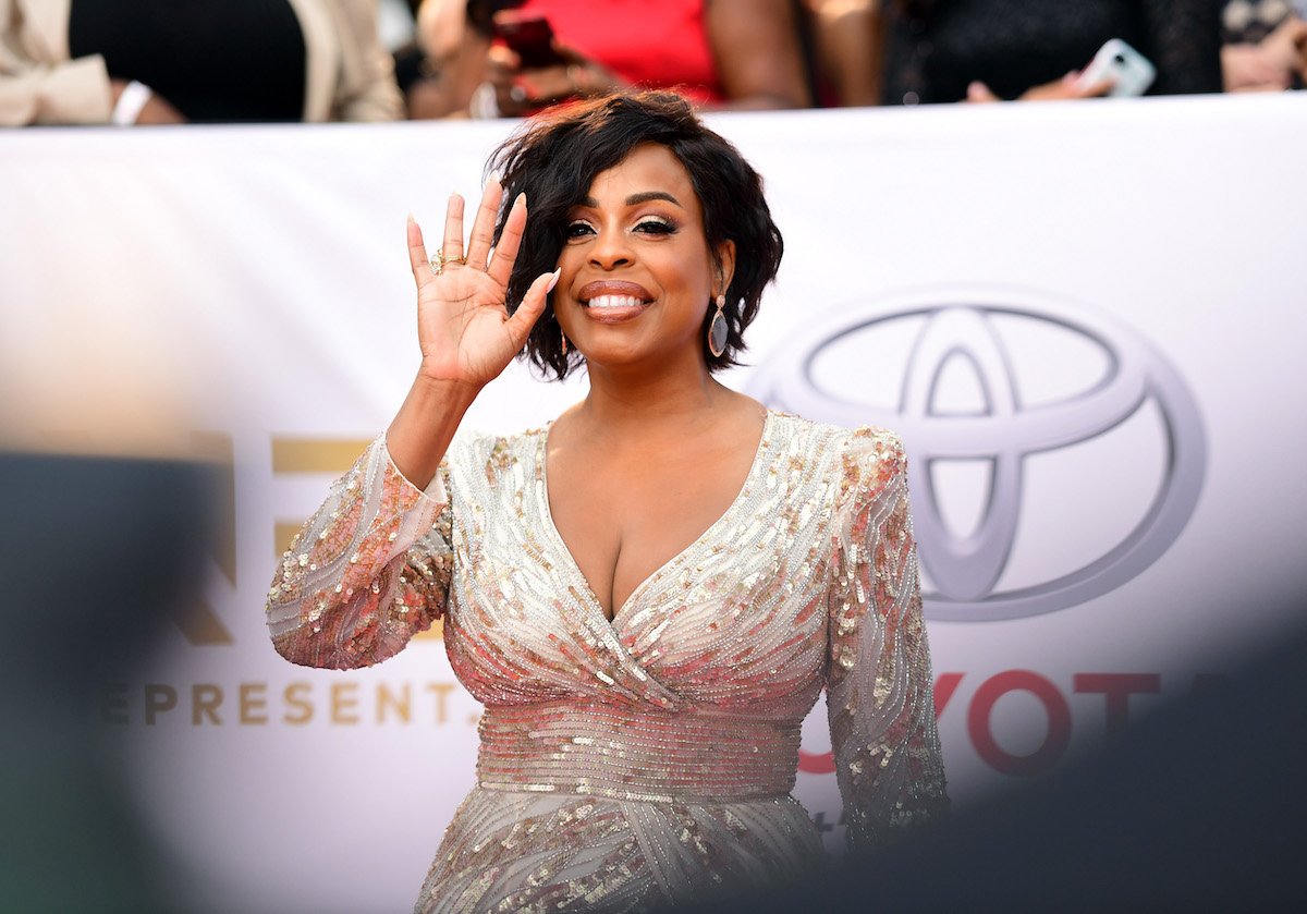 Niecy Nash smiles and waves at the camera in a glittering gold dress