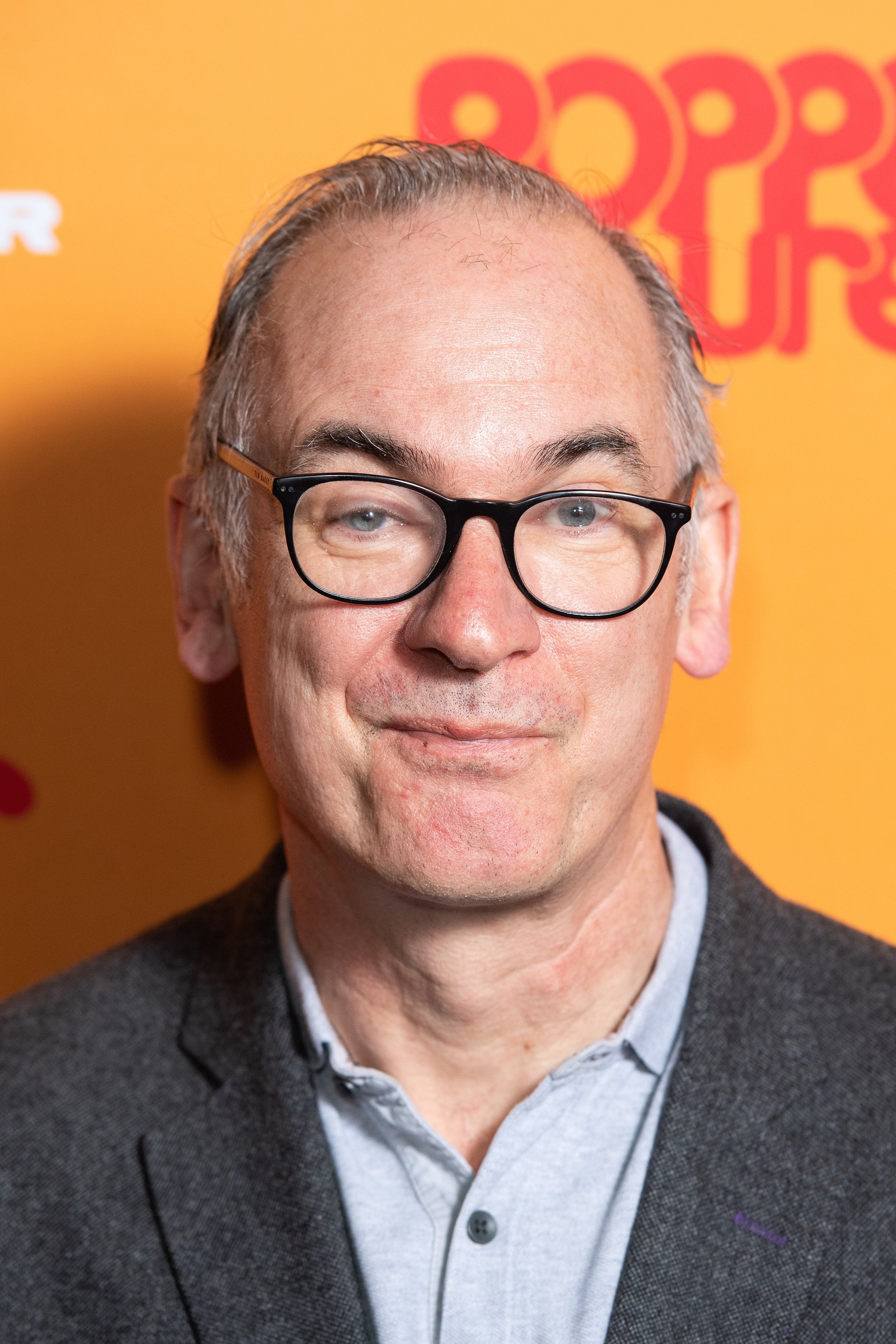 A close-up of actor Paul Ritter with a yellow background behind him