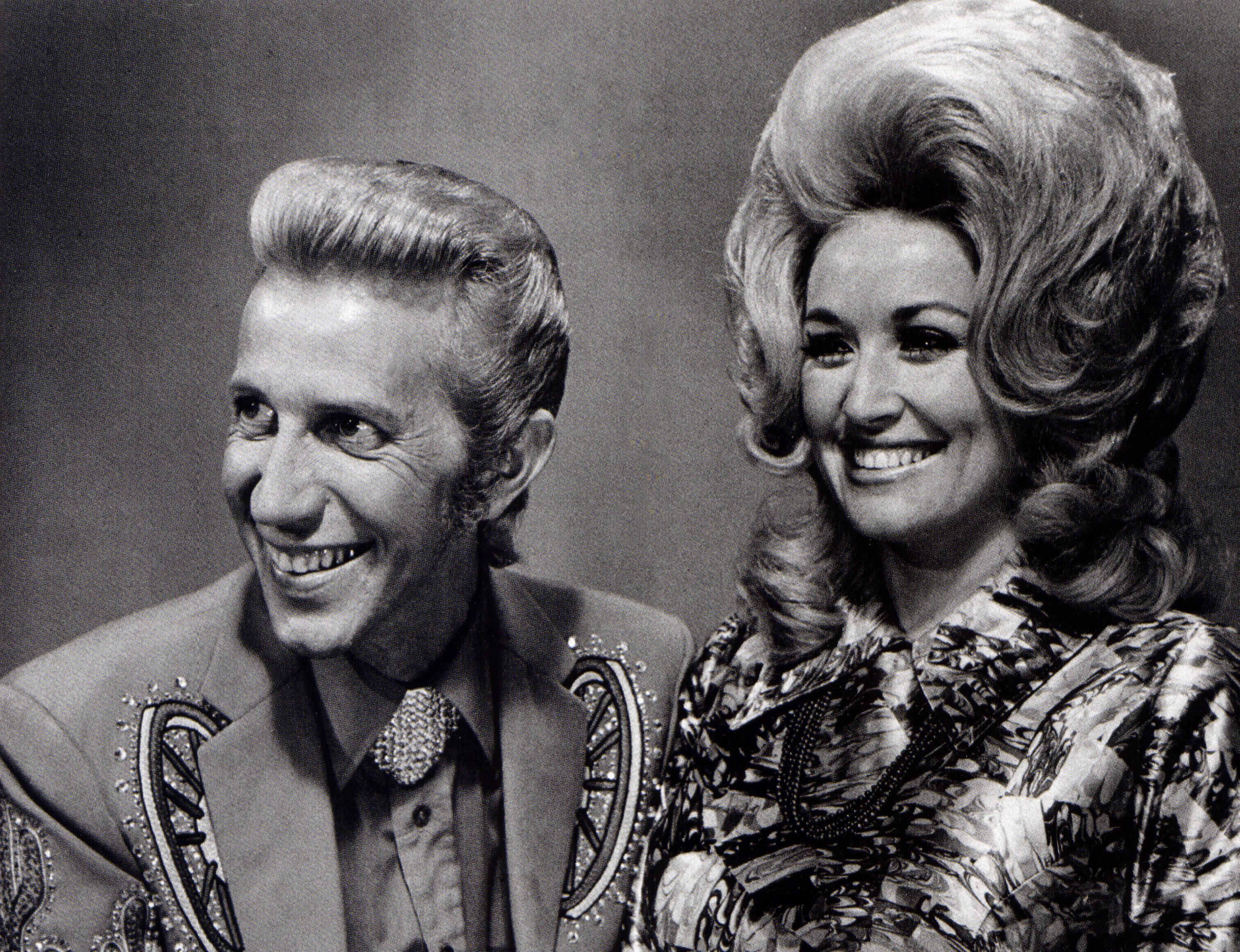 Black and white photo of Porter Wagoner and Dolly Parton. They're smiling looking to the left of the camera.