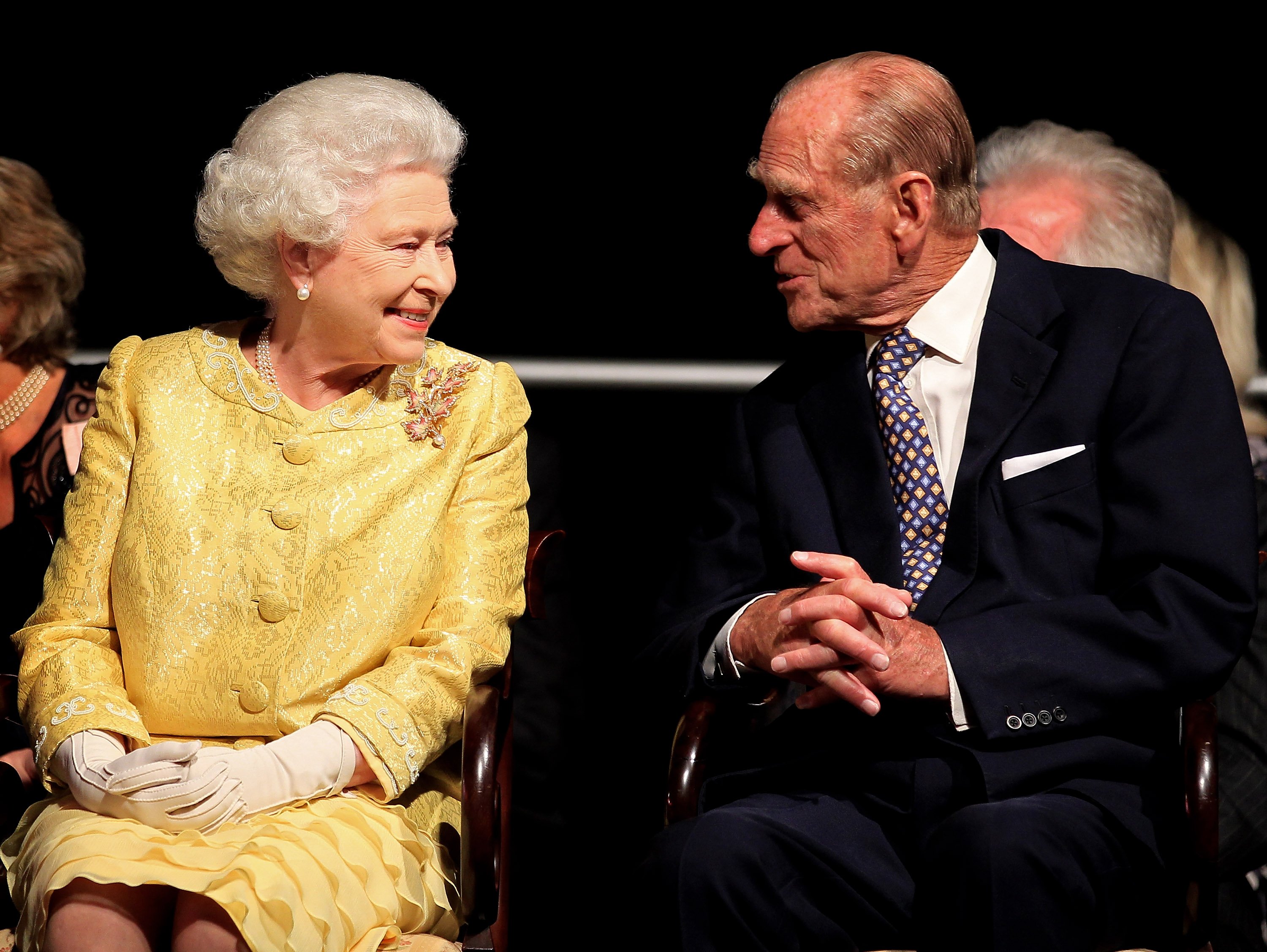 Queen Elizabeth II in a yellow dress and Prince Philip in a suit and tie seated next to each other and chatting at event in Canada