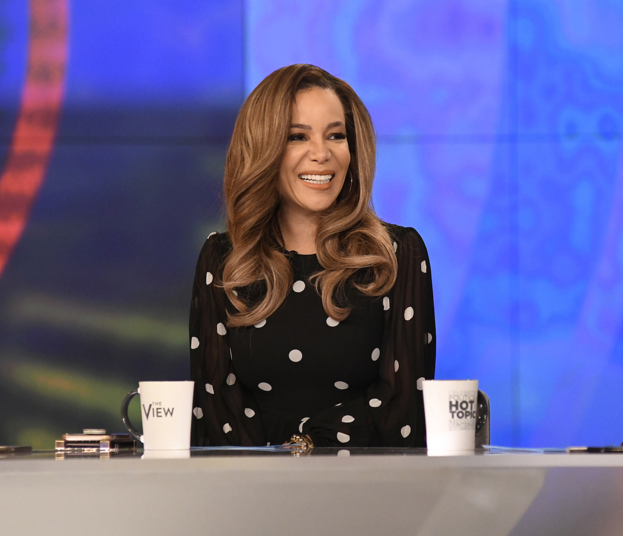 Sunny Hostin in a black top with white dots, smiling at the desk of 'The View'