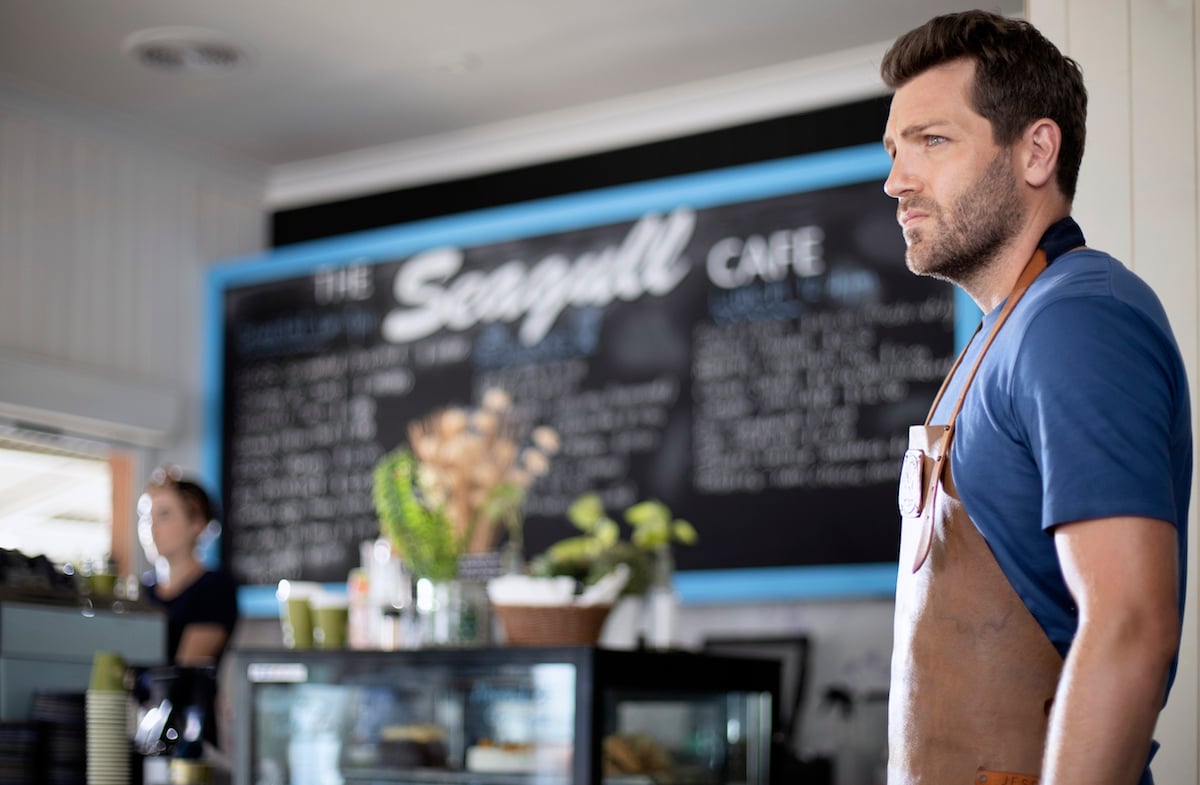 Tim Ross, in profile, standing in front of menu board in scene from Hearts Down Under 