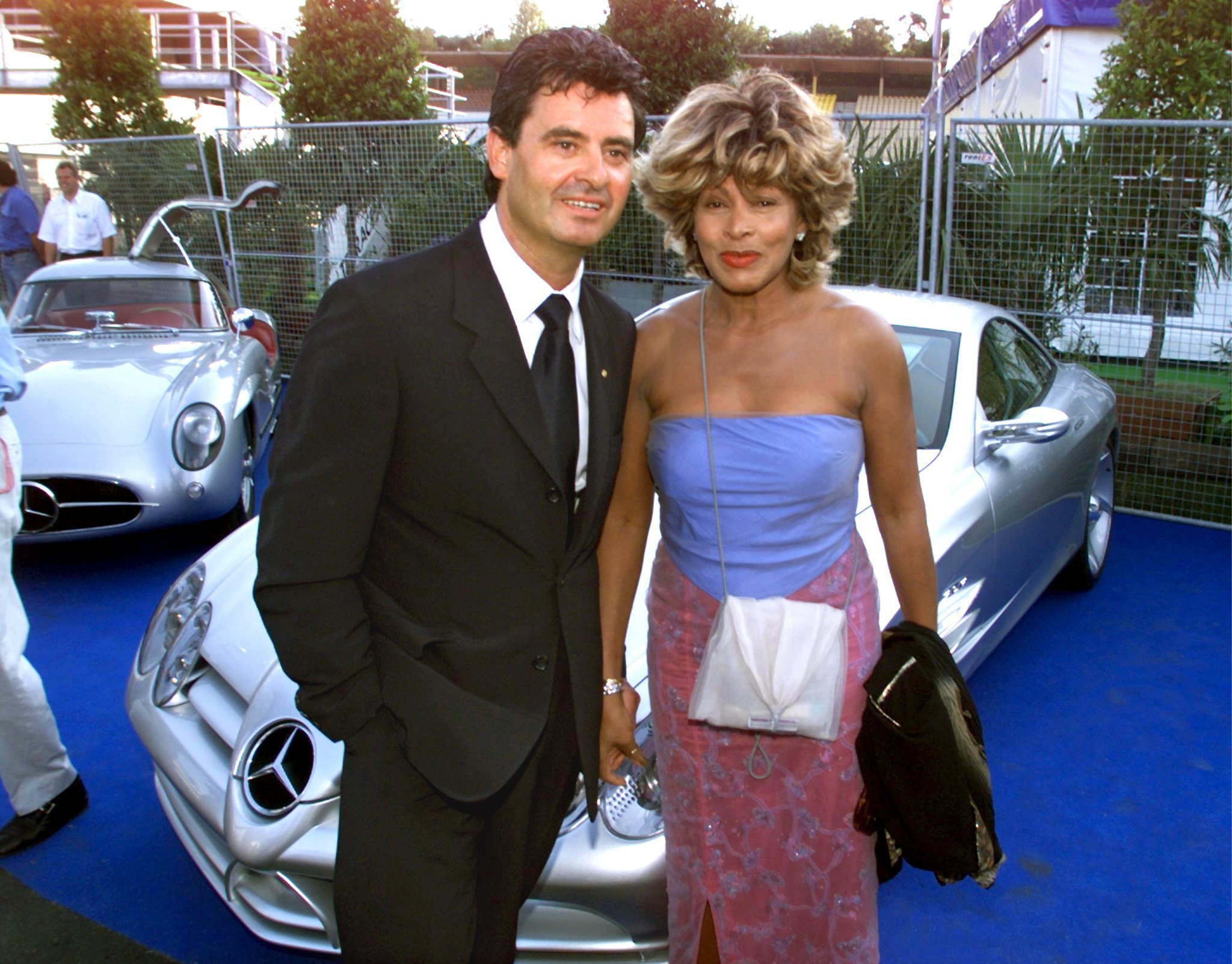 Tina Turner and Erwin Bach stand in a parking lot of fancy cars