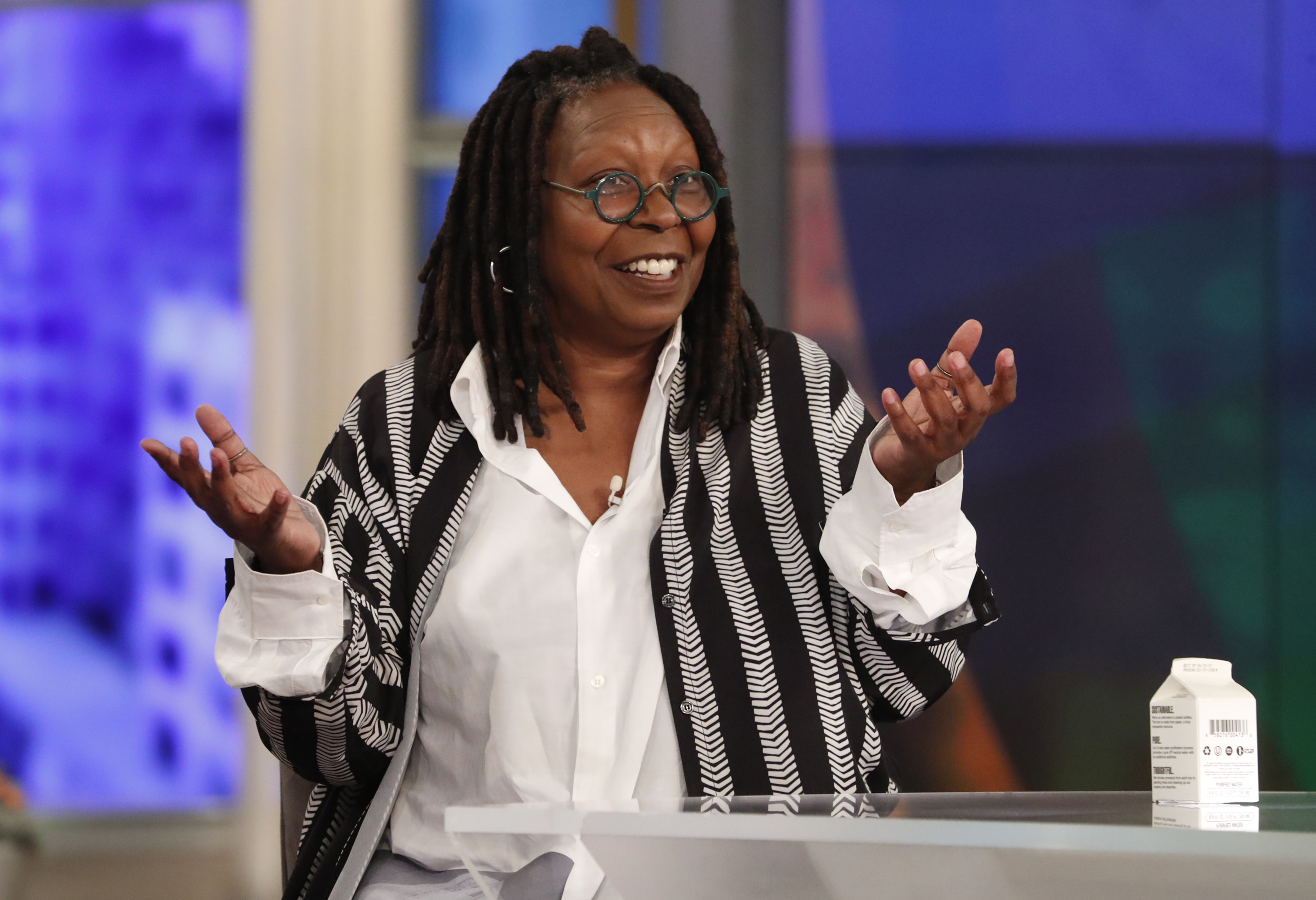 Whoopi Goldberg smiling in a white top and black-and-gray striped vest at the table of 'The View'