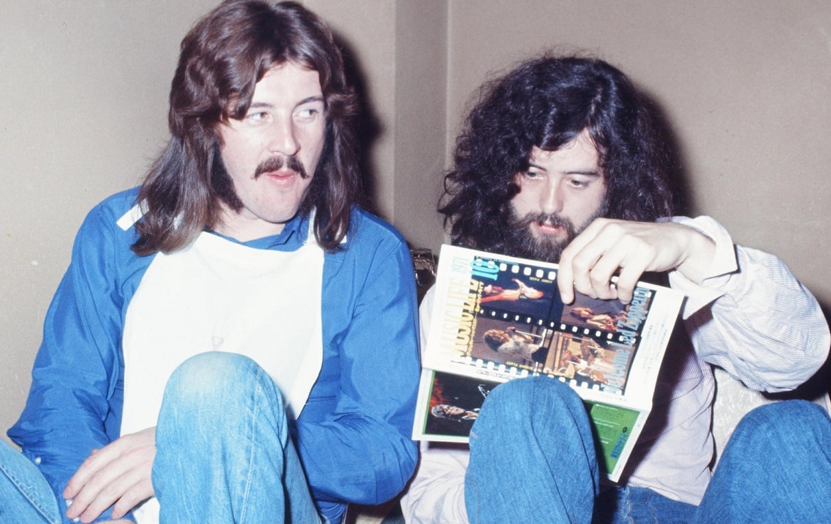 John Bonham looks over at Jimmy Page, who's seated next to him and looking at a magazine