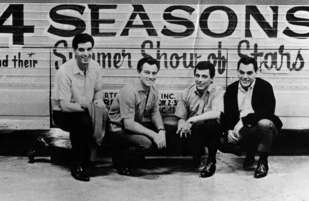 The Four Seasons kneel down at the side of their tour bus and smile for the camera, circa 1964