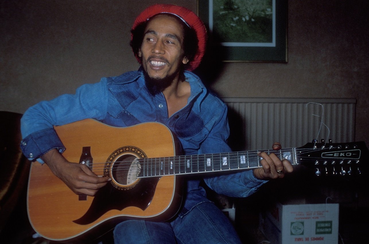 Bob Marley smiles and plays an acoustic guitar in a hotel room in 1978