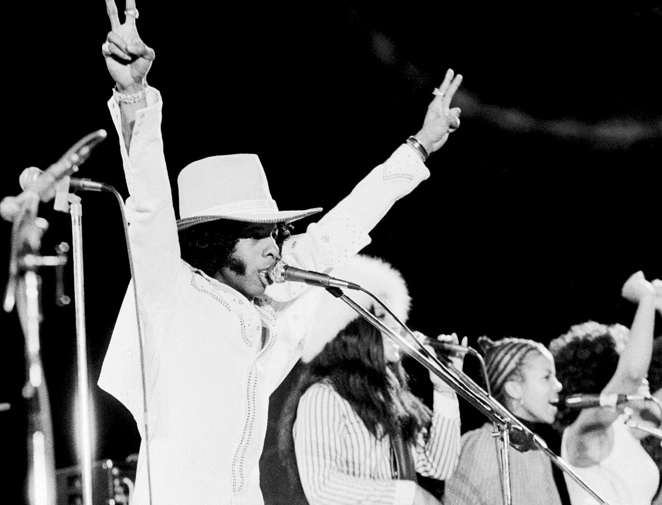 Sly Stone has both arms raised and flashes the peace sign as he sings into a microphone at a concert