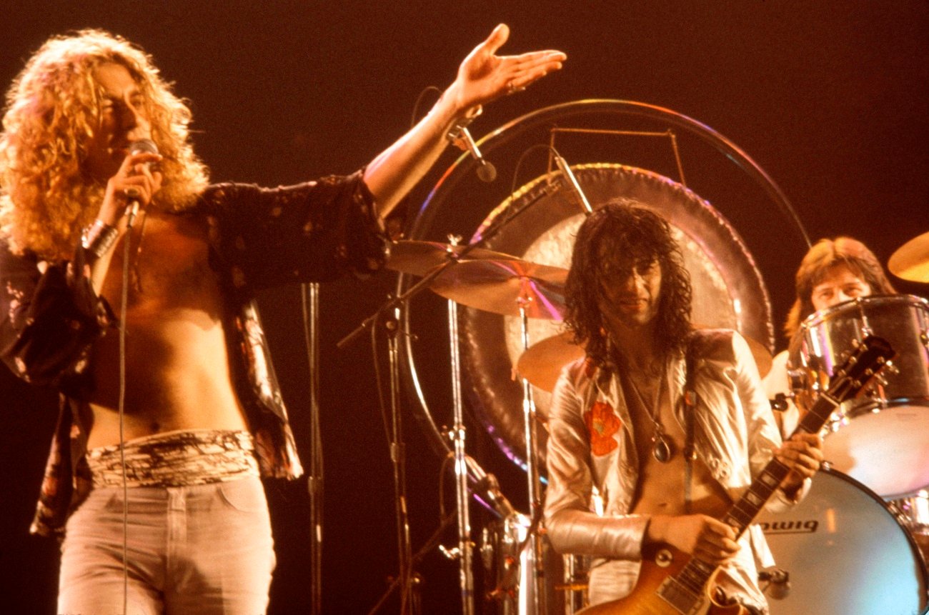 Robert Plant gestures and speaks into the microphone as Jimmy Page and John Bonham look on at a Led Zeppelin show.