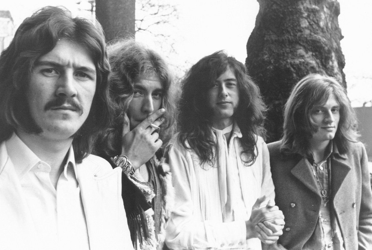 Led Zeppelin band photo, taken outdoors. The members of the group look into the camera and Robert Plant smokes a cigarette