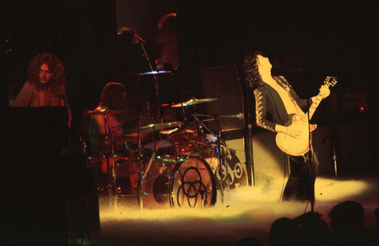 Jimmy Page plays guitar in a spotlight over a sea of smoke with Robert Plant and John Bonham behind him
