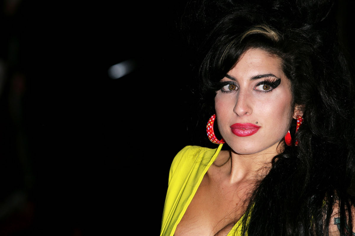 headshot of Amy Winehouse in a yellow top with red earrings against a black background