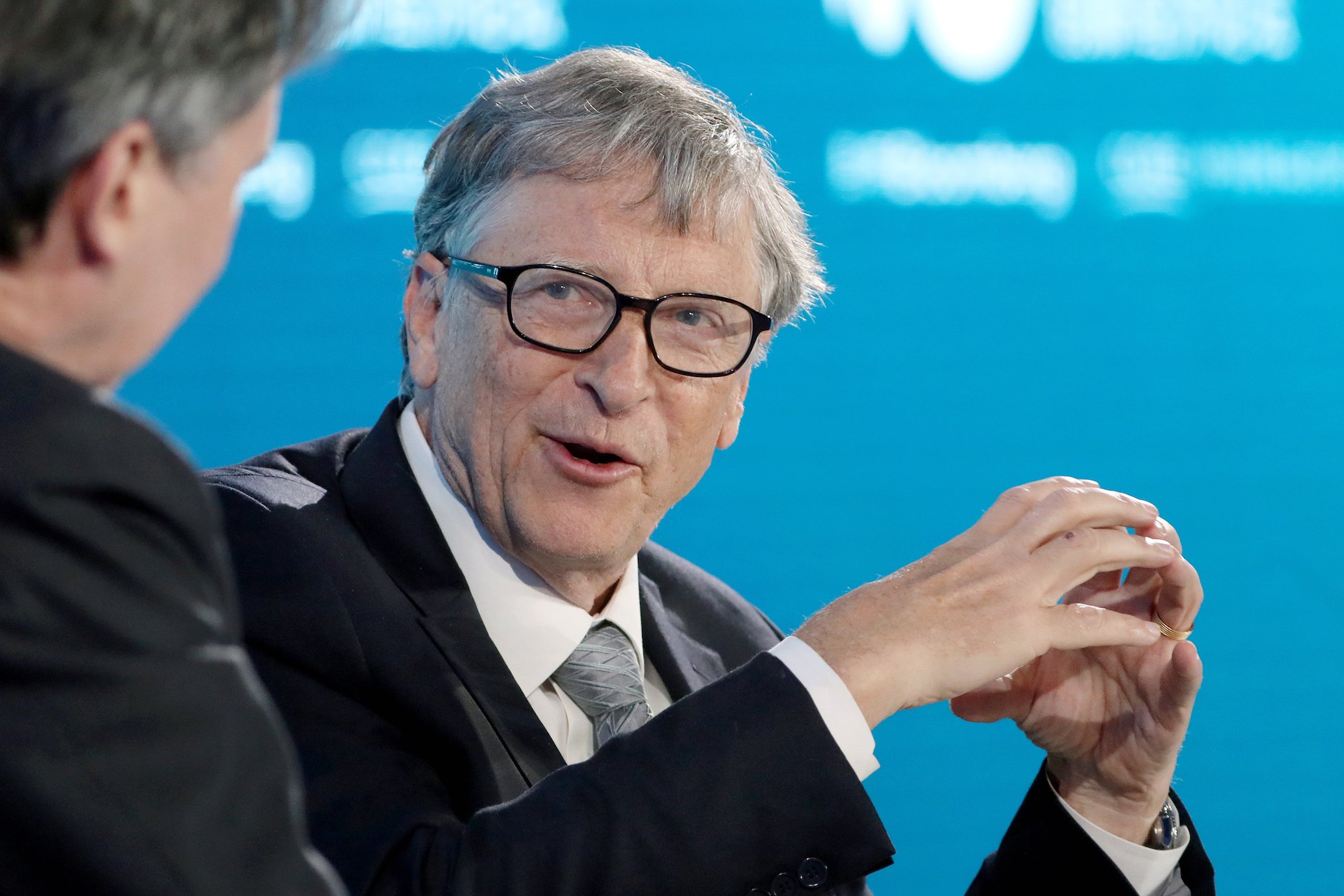 Bill Gates age 64 sitting against a blue background while speaking at a conference