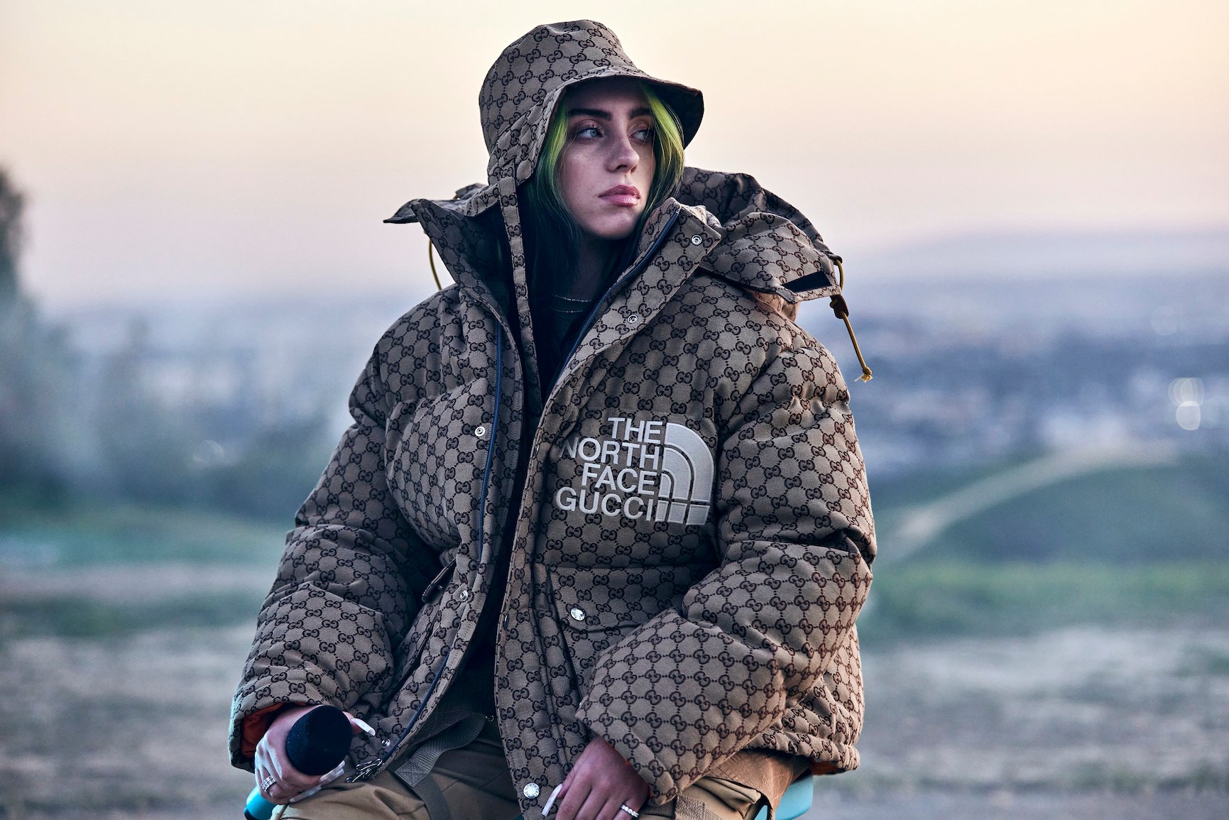 Billie Eilish sitting outside in a puffy plaid jacket with a matching hat