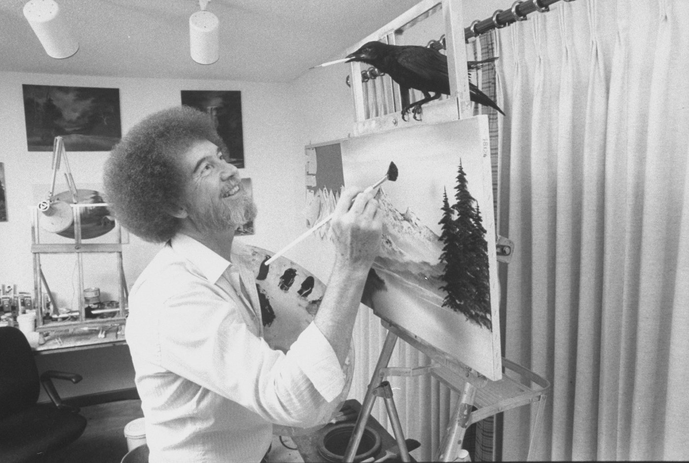 TV painting instructor/artist Bob Ross paints a landscape as his pet crow holds a paintbrush in its beak while perched on Ross's easel