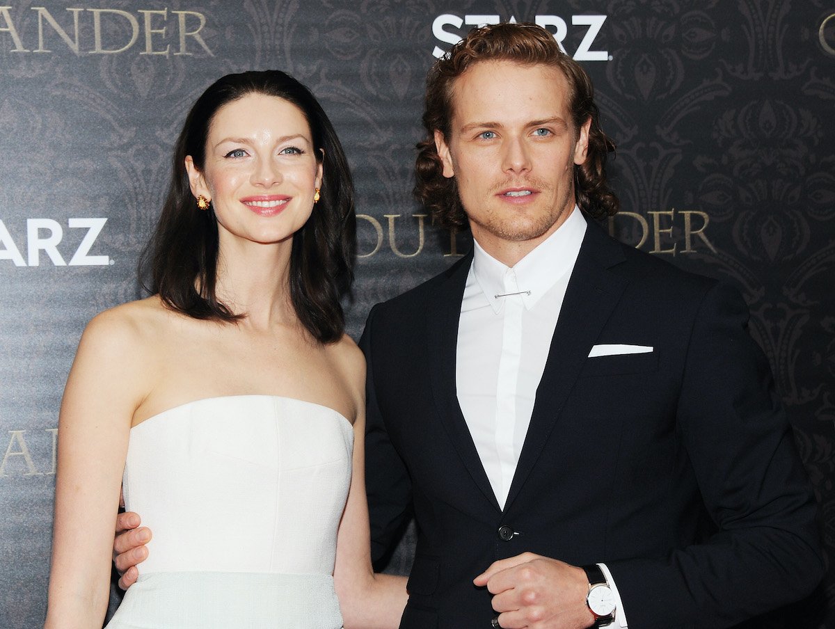 Caitriona Balfe smiles in a strapless white dress and poses next to Sam Heughan, who's wearing a blue suit. They're standing in front of a grey ornate backdrop that says 'Outlander' and 'STARZ.'
