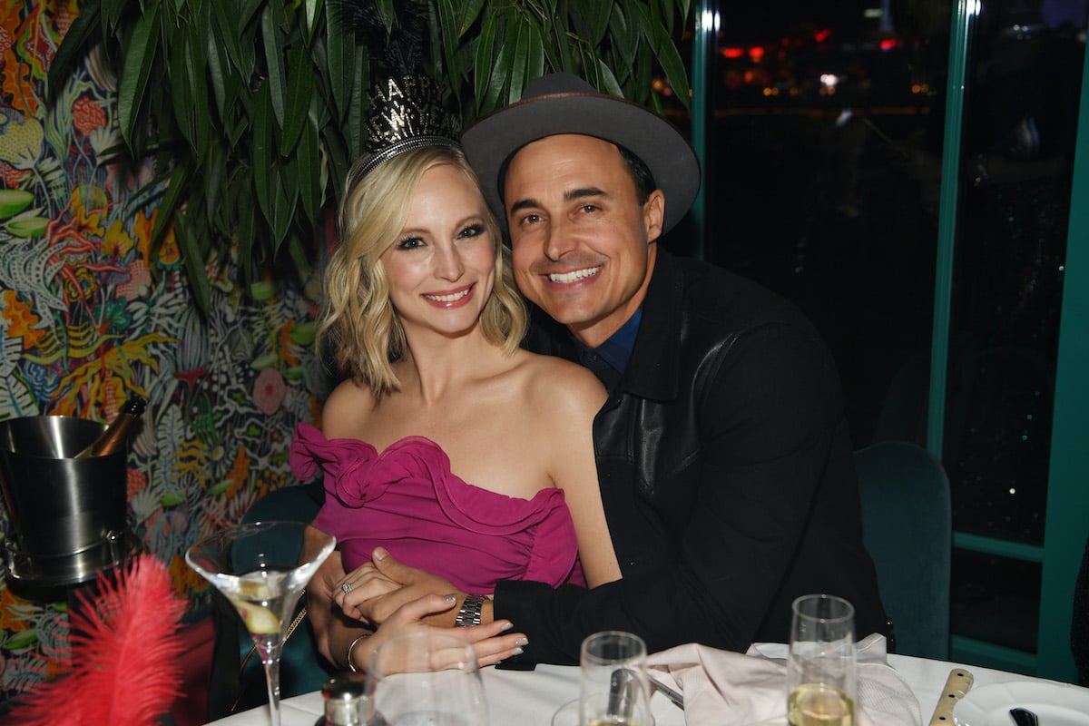 Candice Accola King sits at a table in a pink dress and a Happy New Year crown. Her husband, Joe King, wraps his arms around her and smiles in a blue shirt, black blazer, and grey hat.