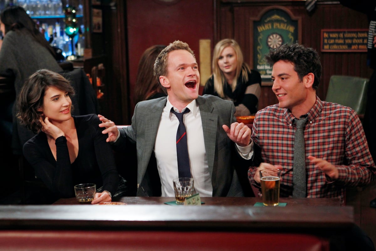 Neil Patrick Harris smiles and gestures wildly, while Cobie Smulders (left) and Josh Radnor (right) look on in a scene from 'How I Met Your Mother'