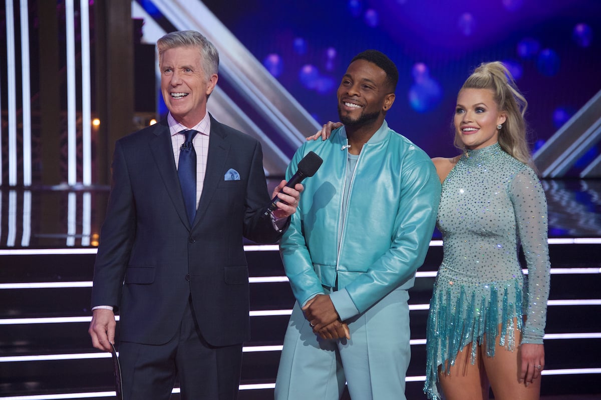 Tom Bergeron holds a microphone for Kel MitchellI, who is standing next to Witney Carson on the DWTS stage