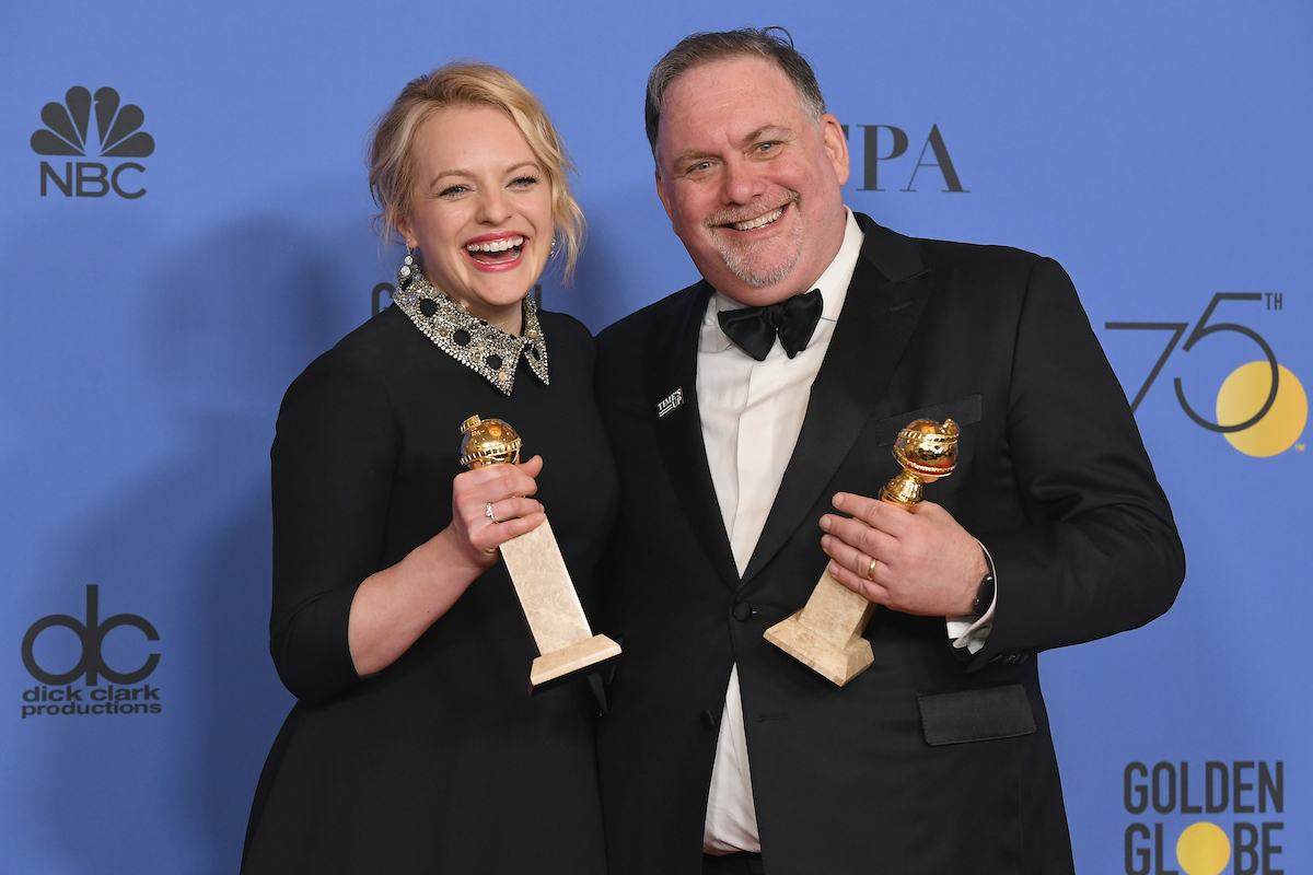 Elisabeth Moss smiling in a black dress and Bruce Miller smiling in a black suit, each holding a Golden Globe trophy at the 2018 Golden Globes