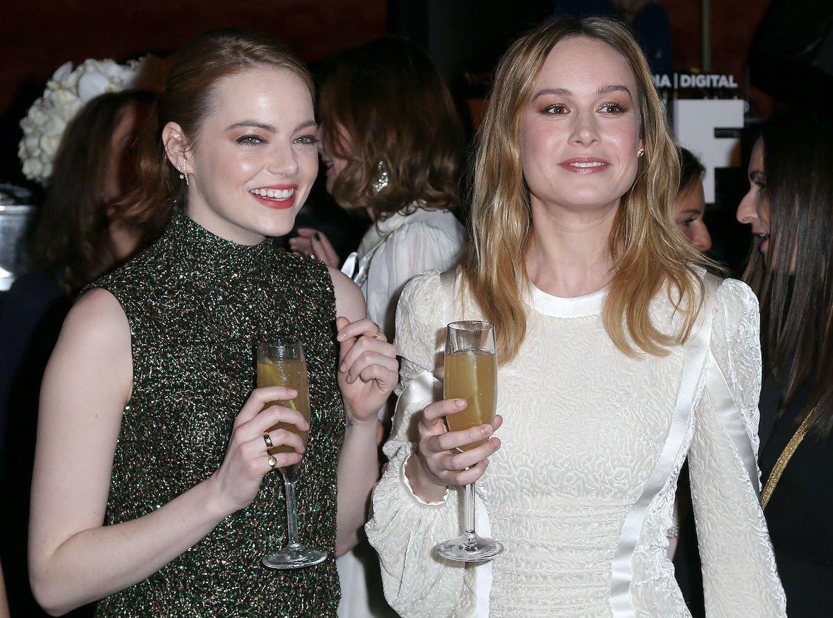 Emma Stone and Brie Larson smile and hold champagne glasses in dresses at an event