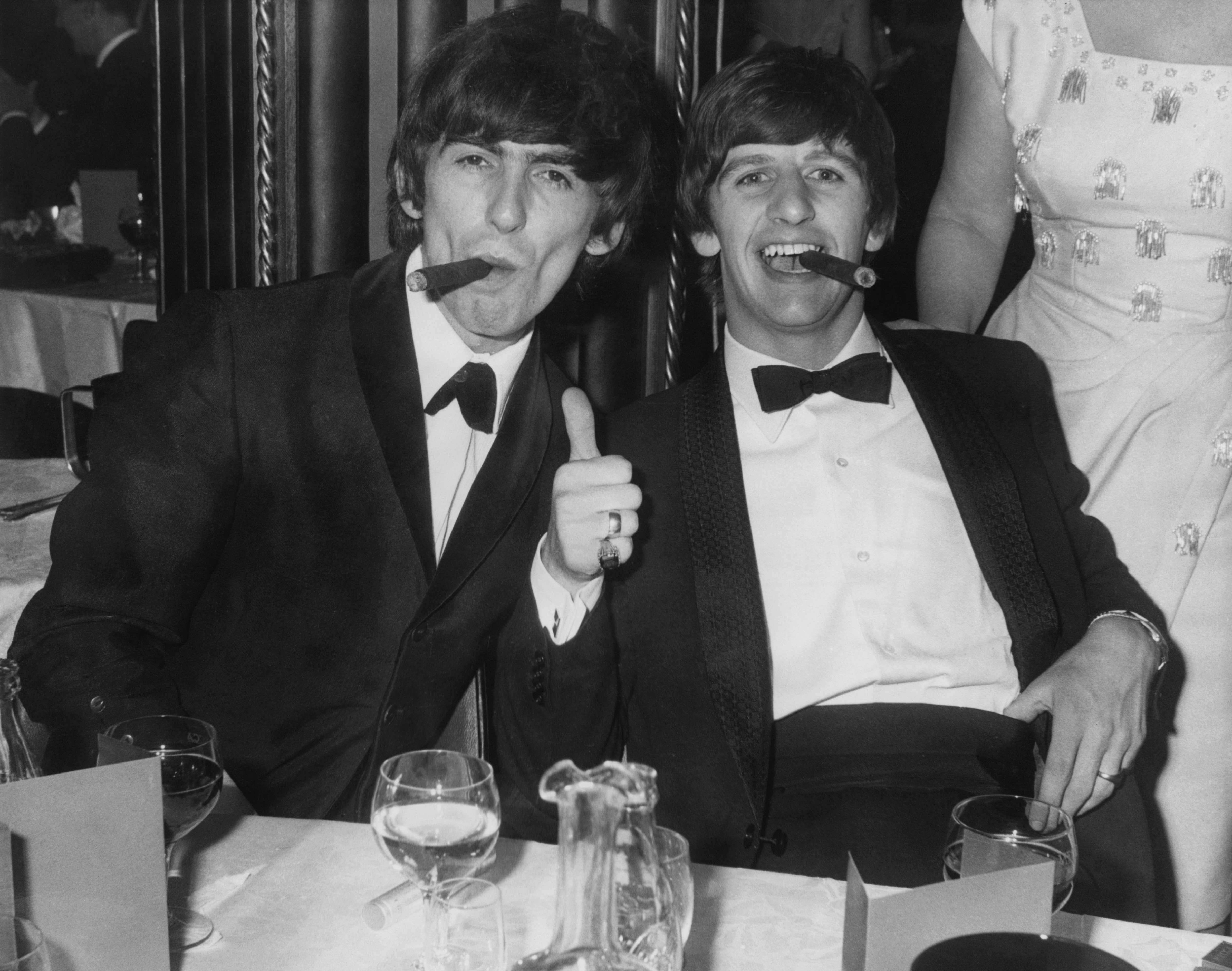 Beatles band mates George Harrison and Ringo Starr sit together during a 1964 awards show. Both are dressed in tuxedos and smoking cigars, while Starr gives a thumbs up.