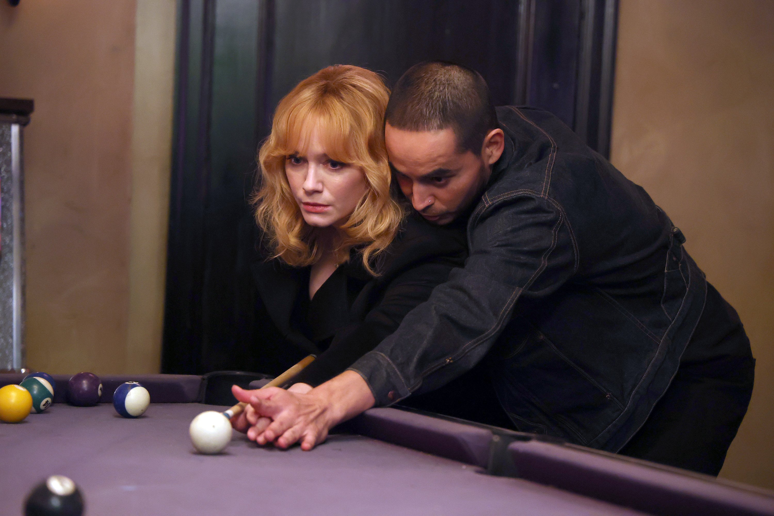 Christina Hendricks and Manny Montana wearing all-black and playing pool as Beth and Rio